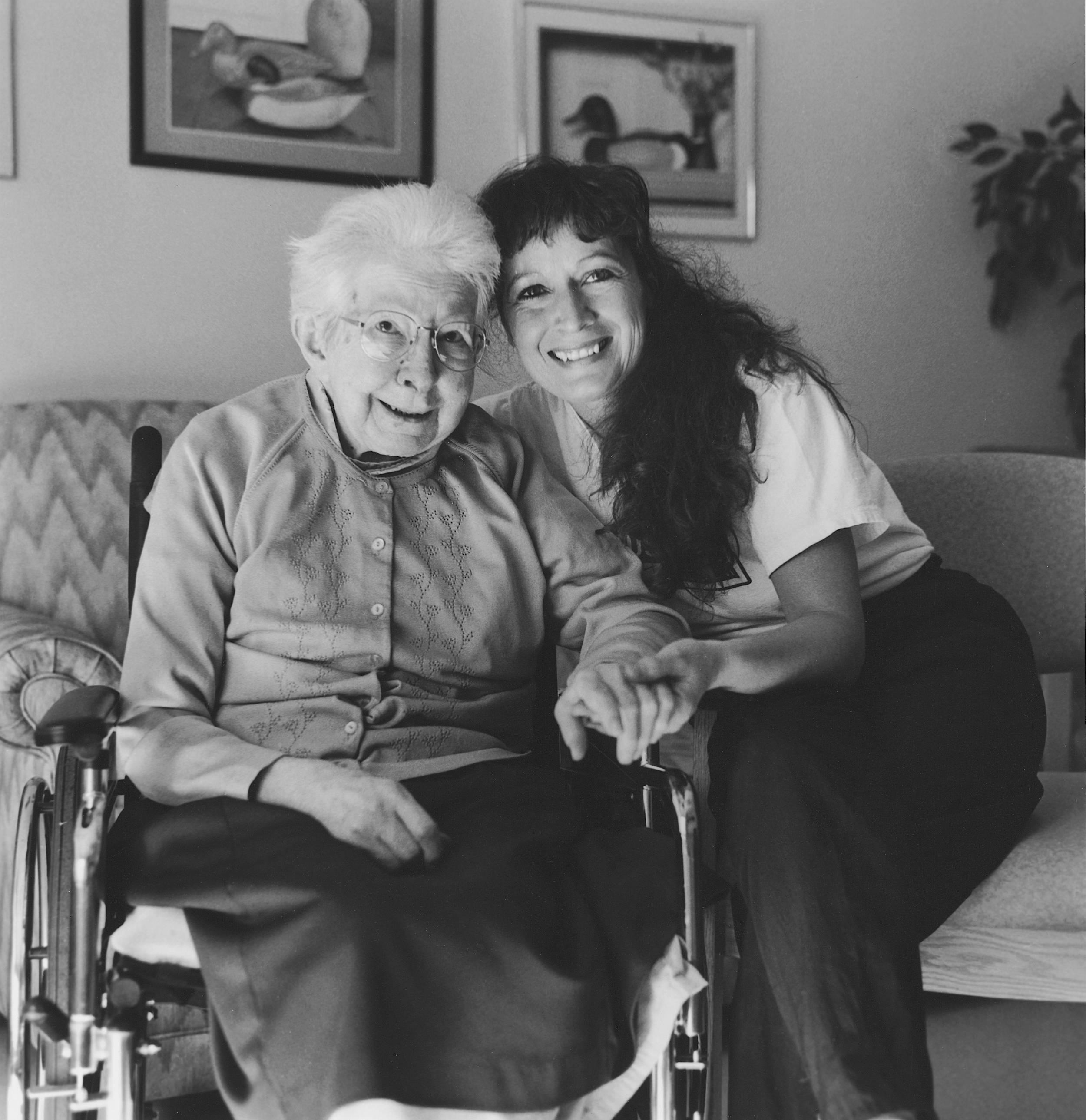 Black and white photo of elderly woman in wheelchair holding the hand of younger woman, both looking up with beautiful smiles in soft morning light.