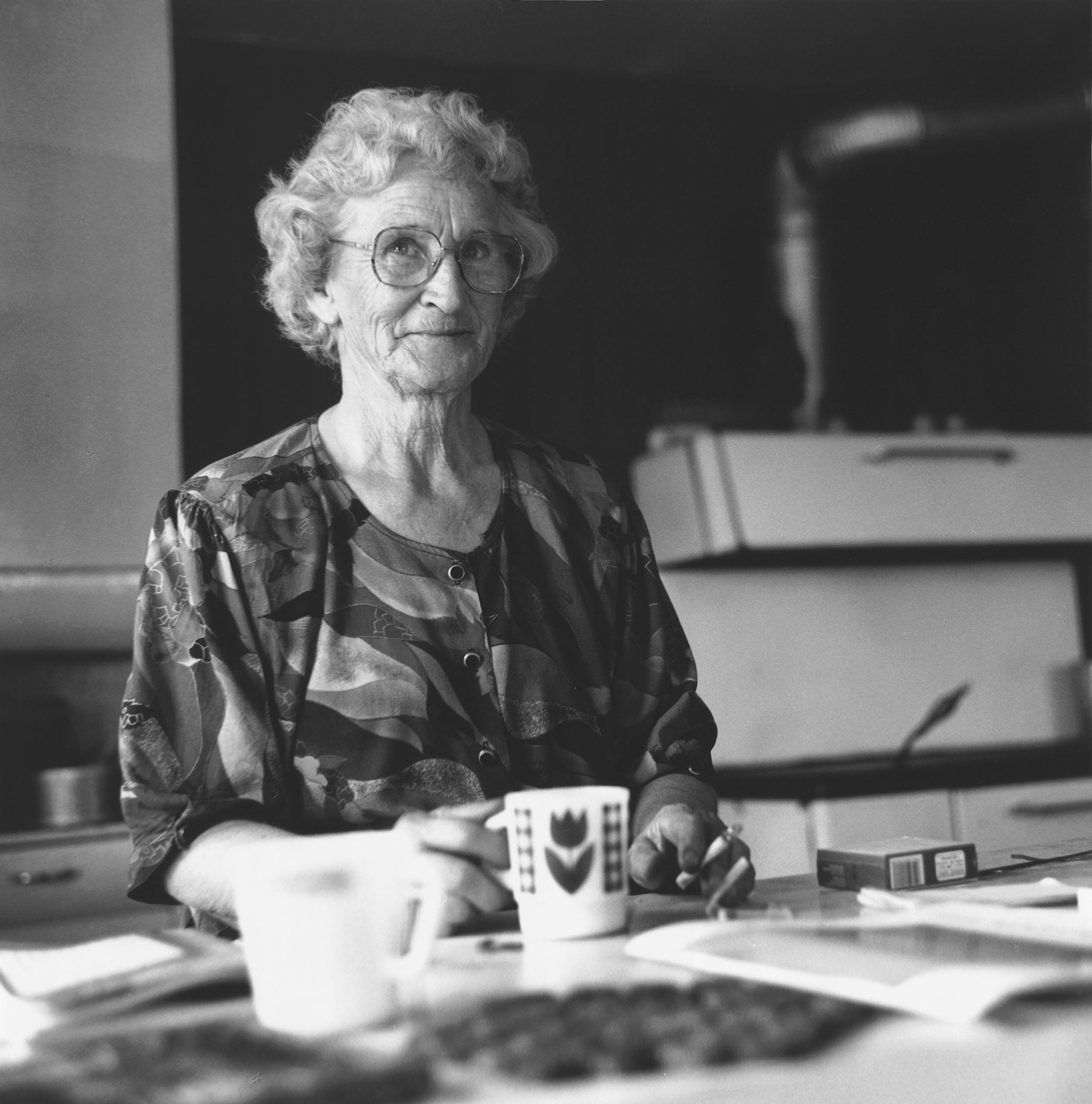 Black and white photo of elderly woman sitting at a table smiling. There are papers and coffee mugs on the table.