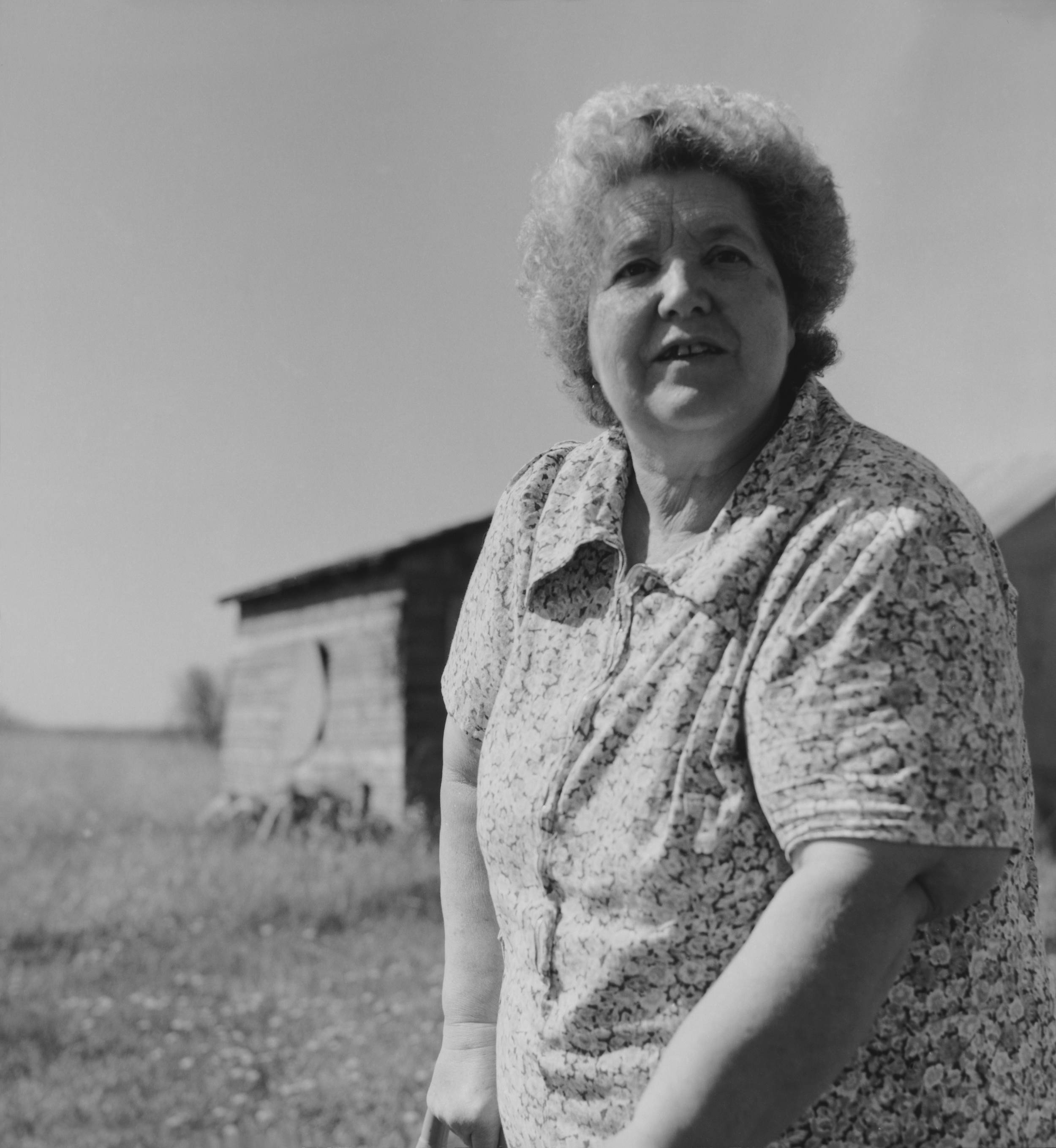 Black and white photo of woman standing in front of old barn out in the prairie. She is wearing a pattern dress.