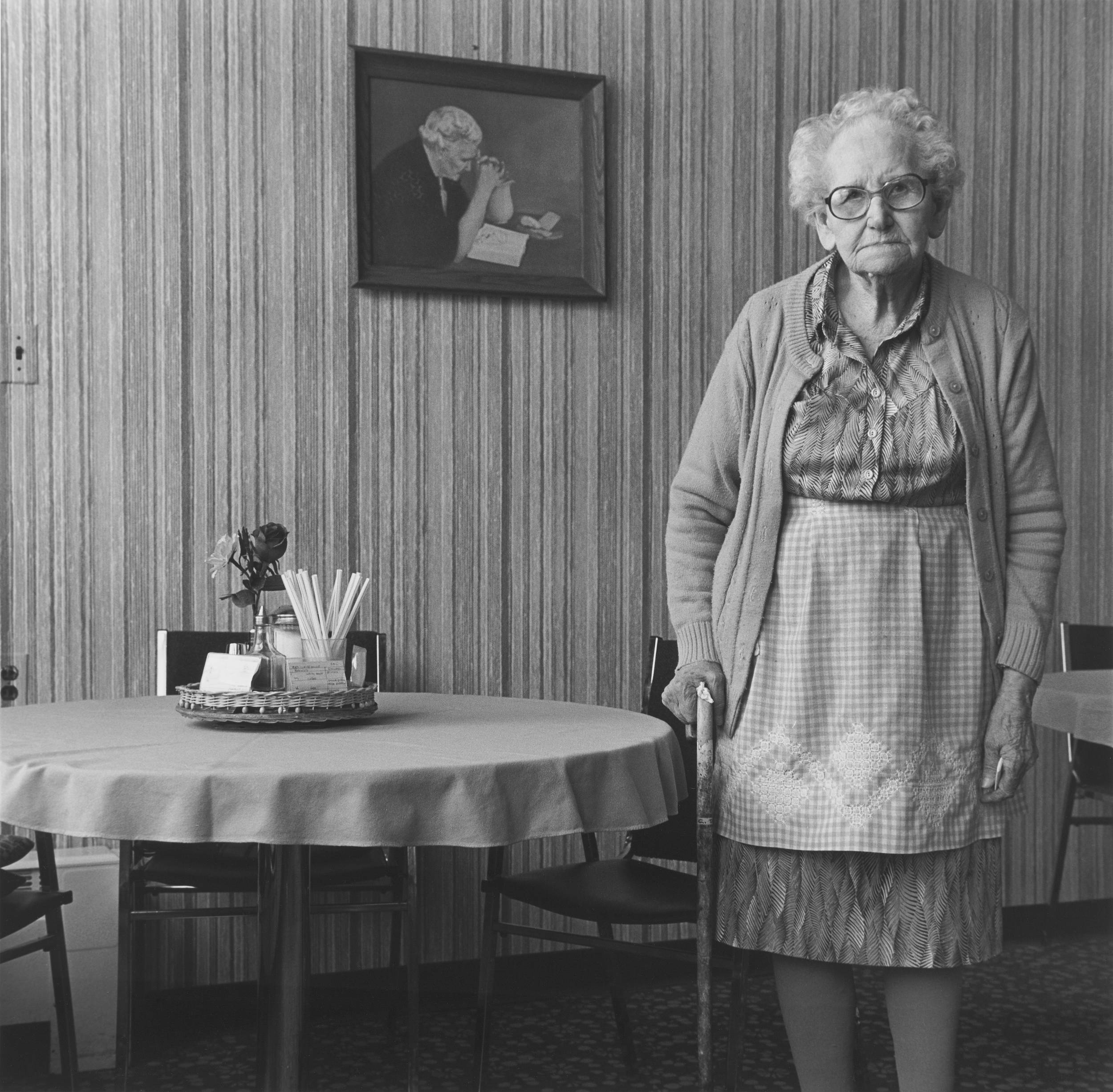 Black and white photo of elderly woman with cane standing beside small dining table, striped wallpaper background.