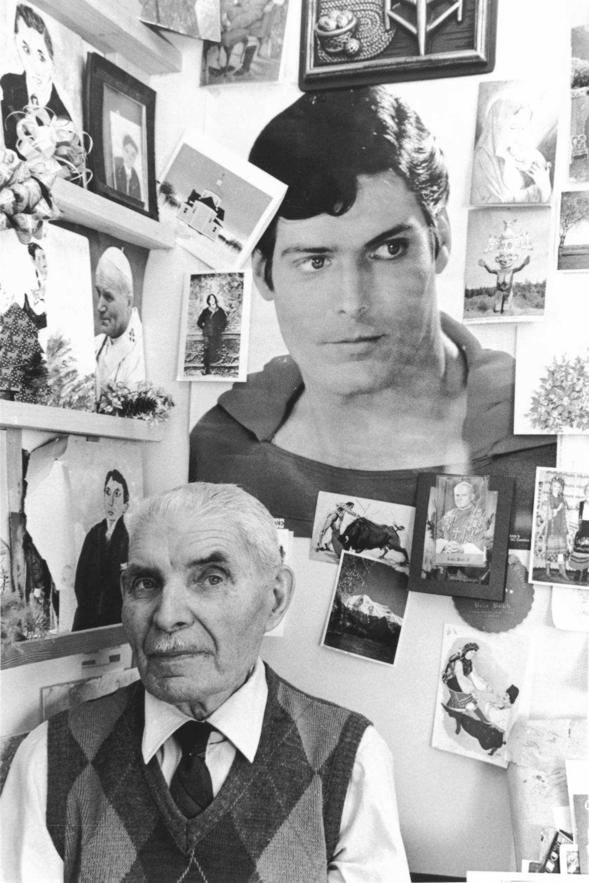 Black and white photo of elderly man, head and shoulders only. Behind him is a large picture of Superman and about a dozen other small paintings.
