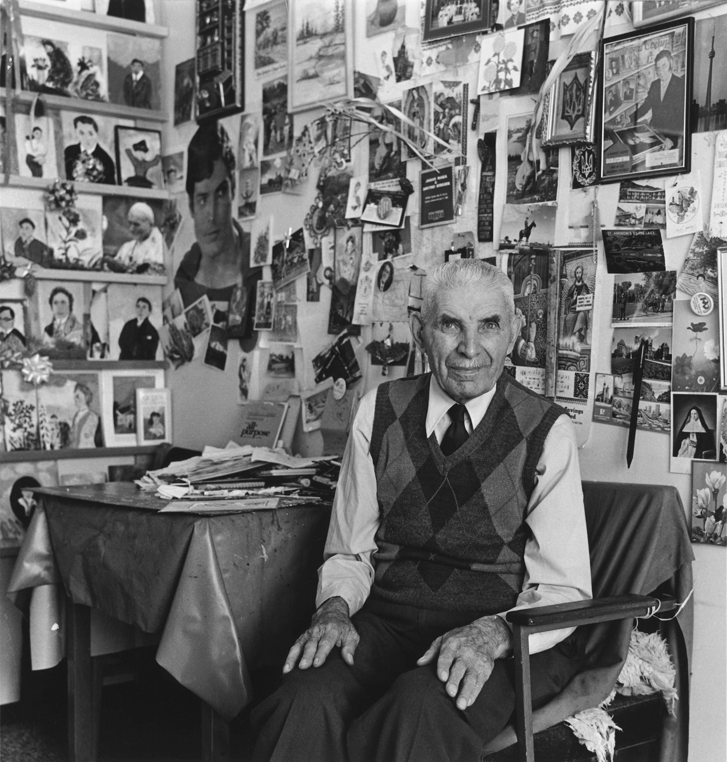 Black and white photo of elderly man seated at home in front of dozens and dozens of small painted pictures behind him. His expression is one of quiet confidence.