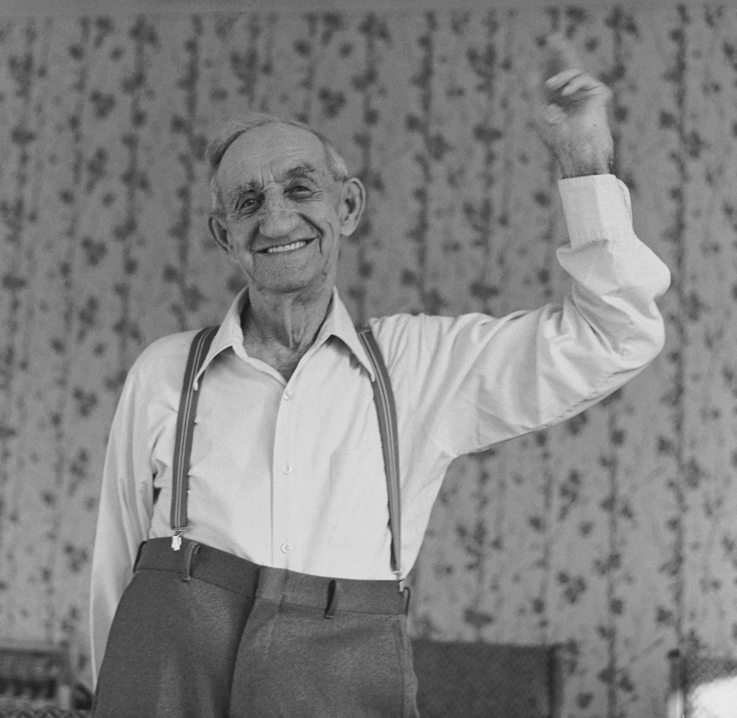 Black and white photo of smiling elderly man, waist up, holding up his right arm above head.