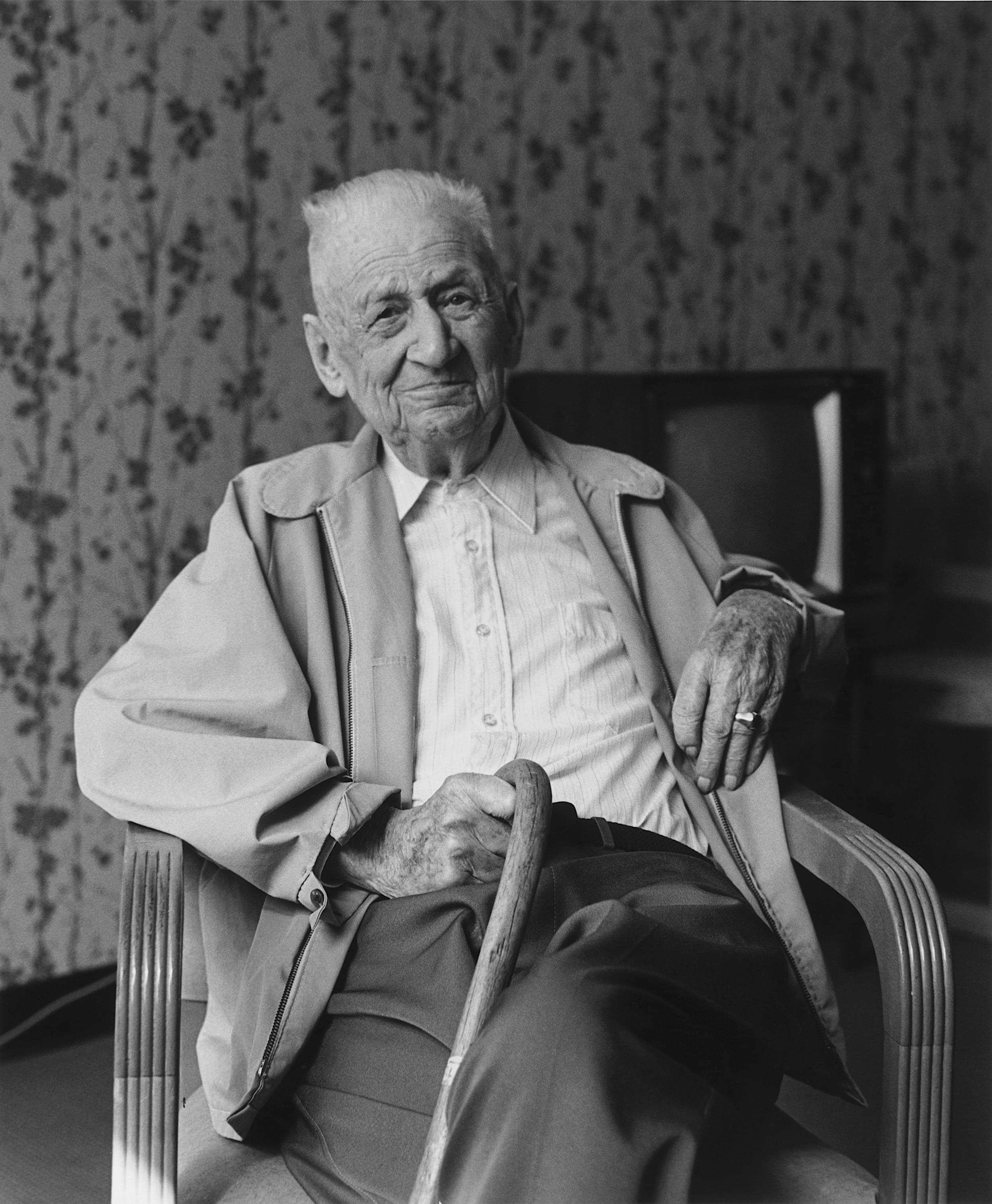 Black and white photo of elderly man seated holding cane in right hand over his lap.