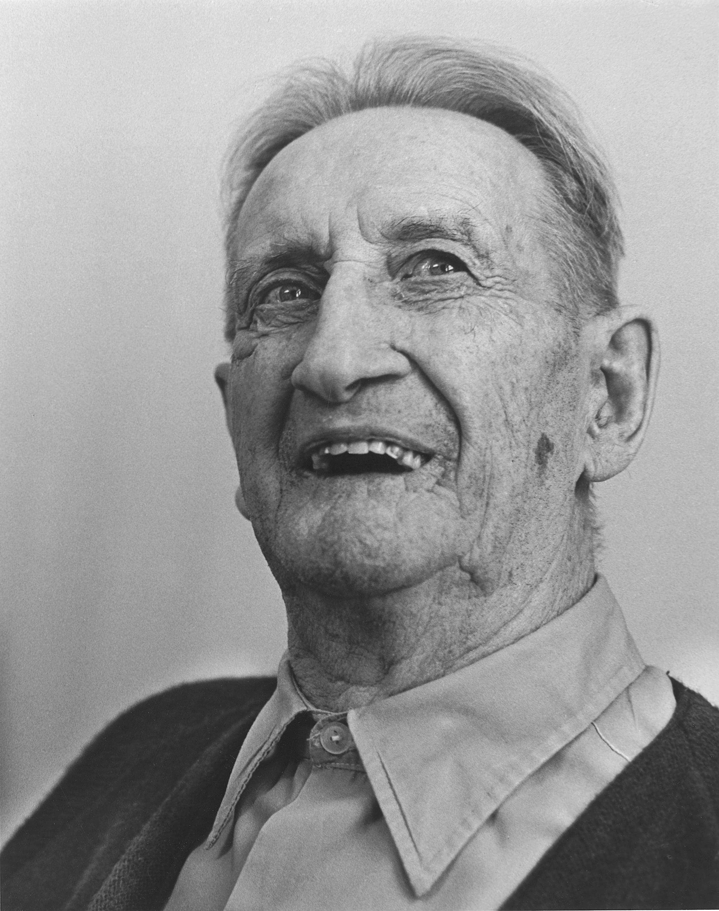 Black and white close-up photo of elderly man wearing shirt with top button tied up.