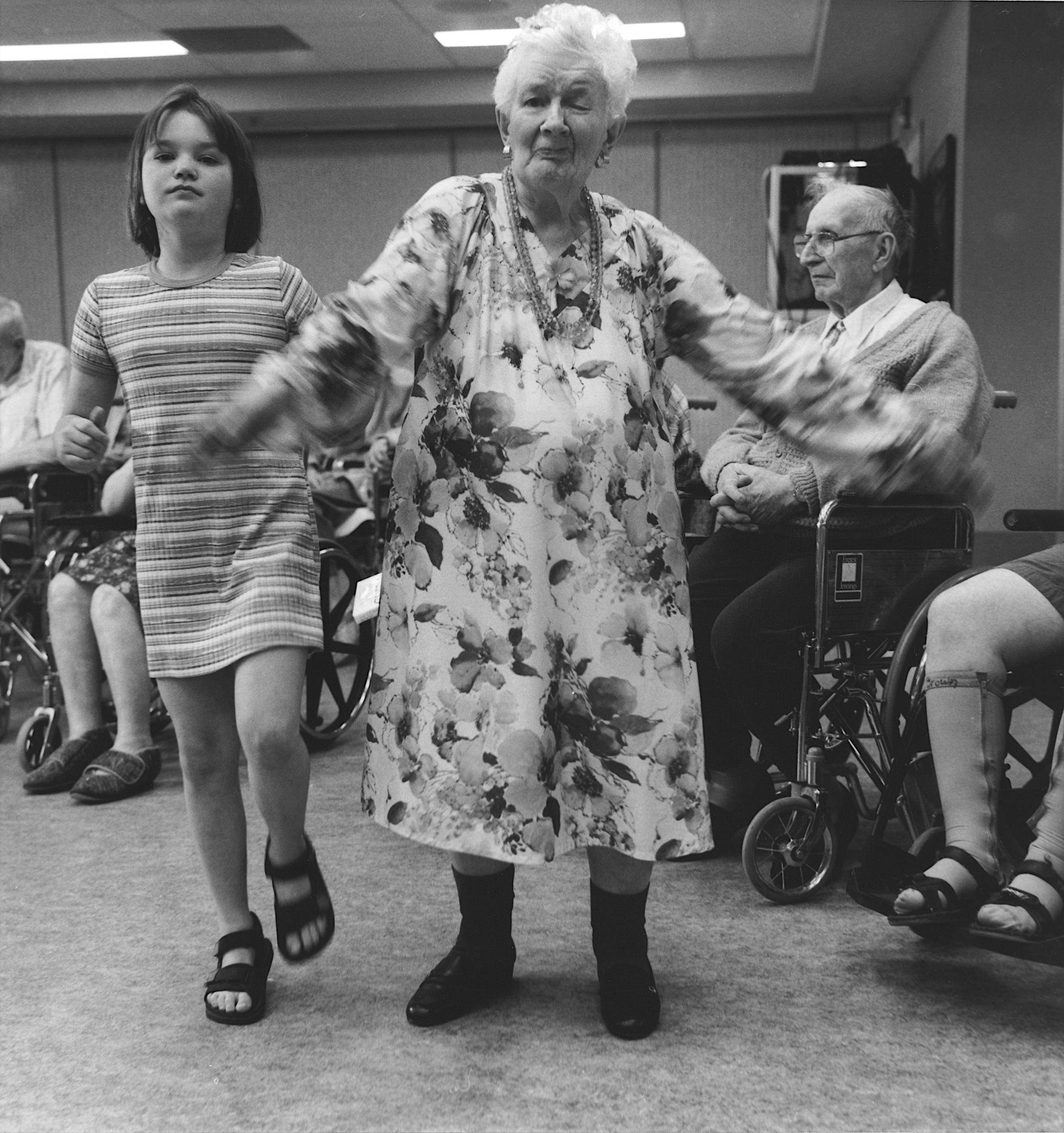 Black and white photo of elderly woman wearing beautiful patterned dress dancing beside young girl with two people in wheelchairs in background.