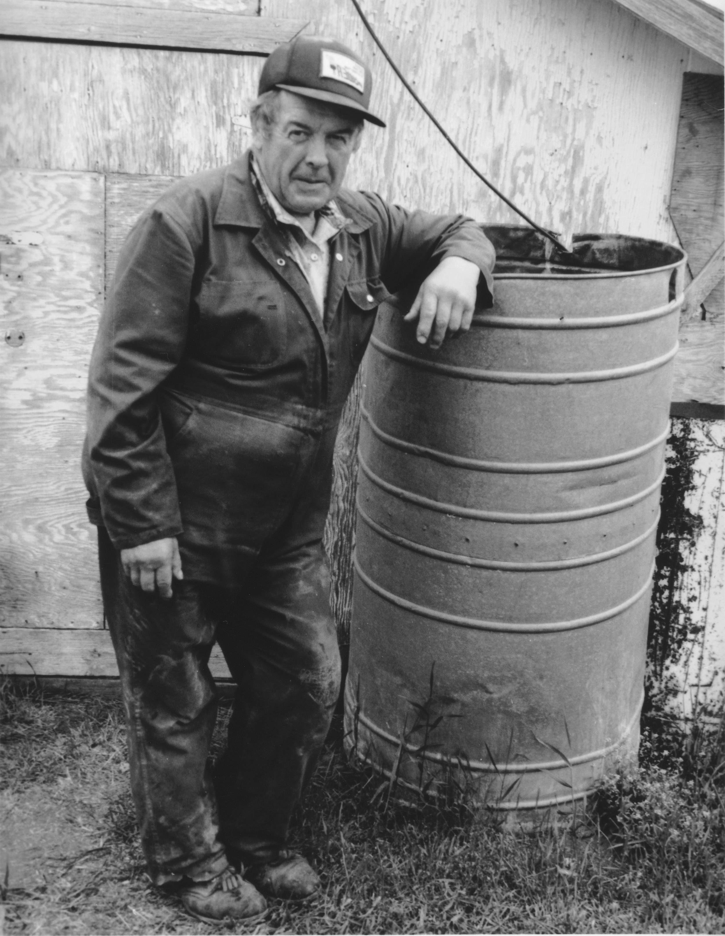 Black and white photo of elderly man leaning against an old metal barrel.