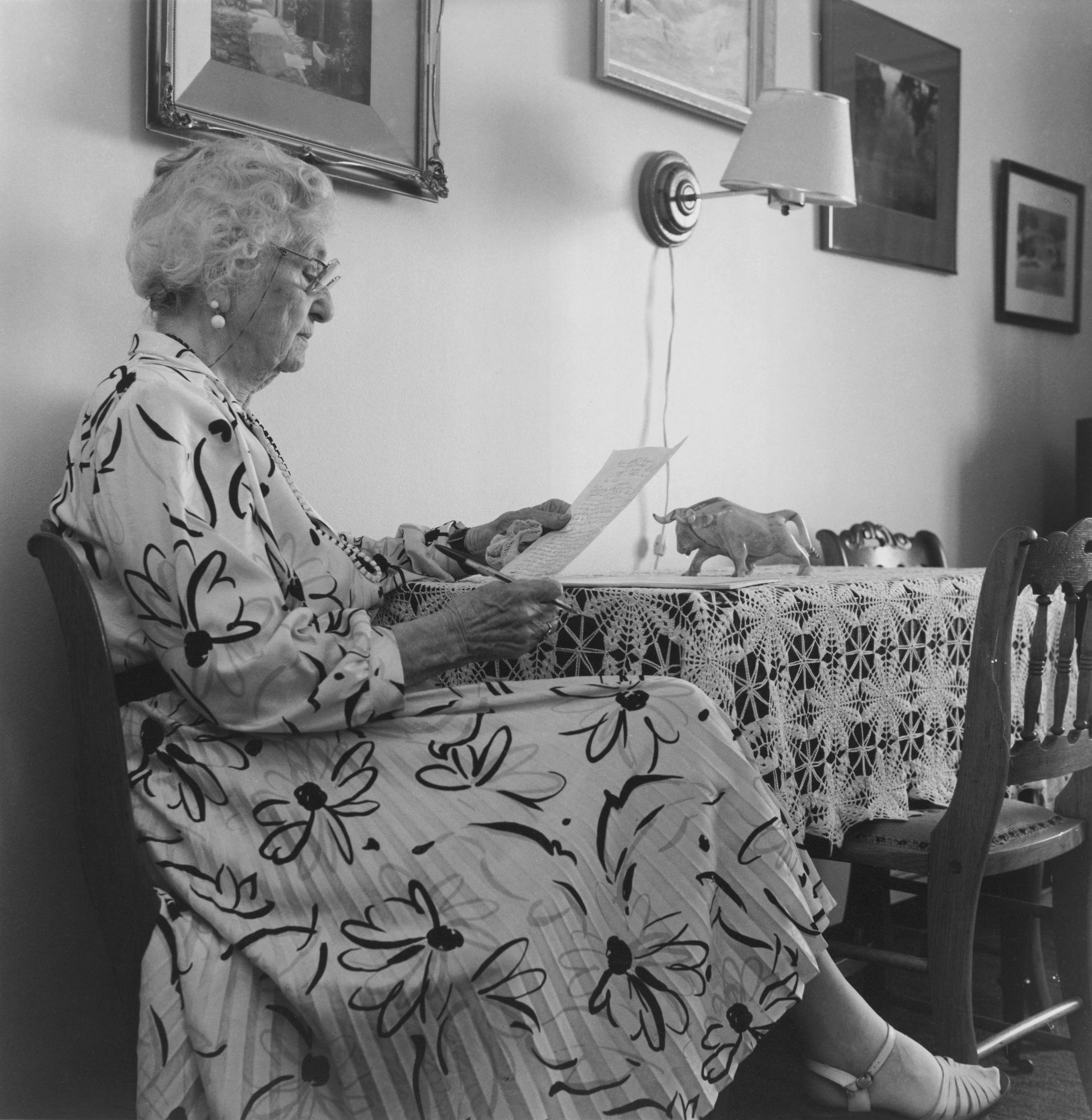 Black and white photo of elderly woman wearing long patterned dress seated sideways at end of table reading letter.