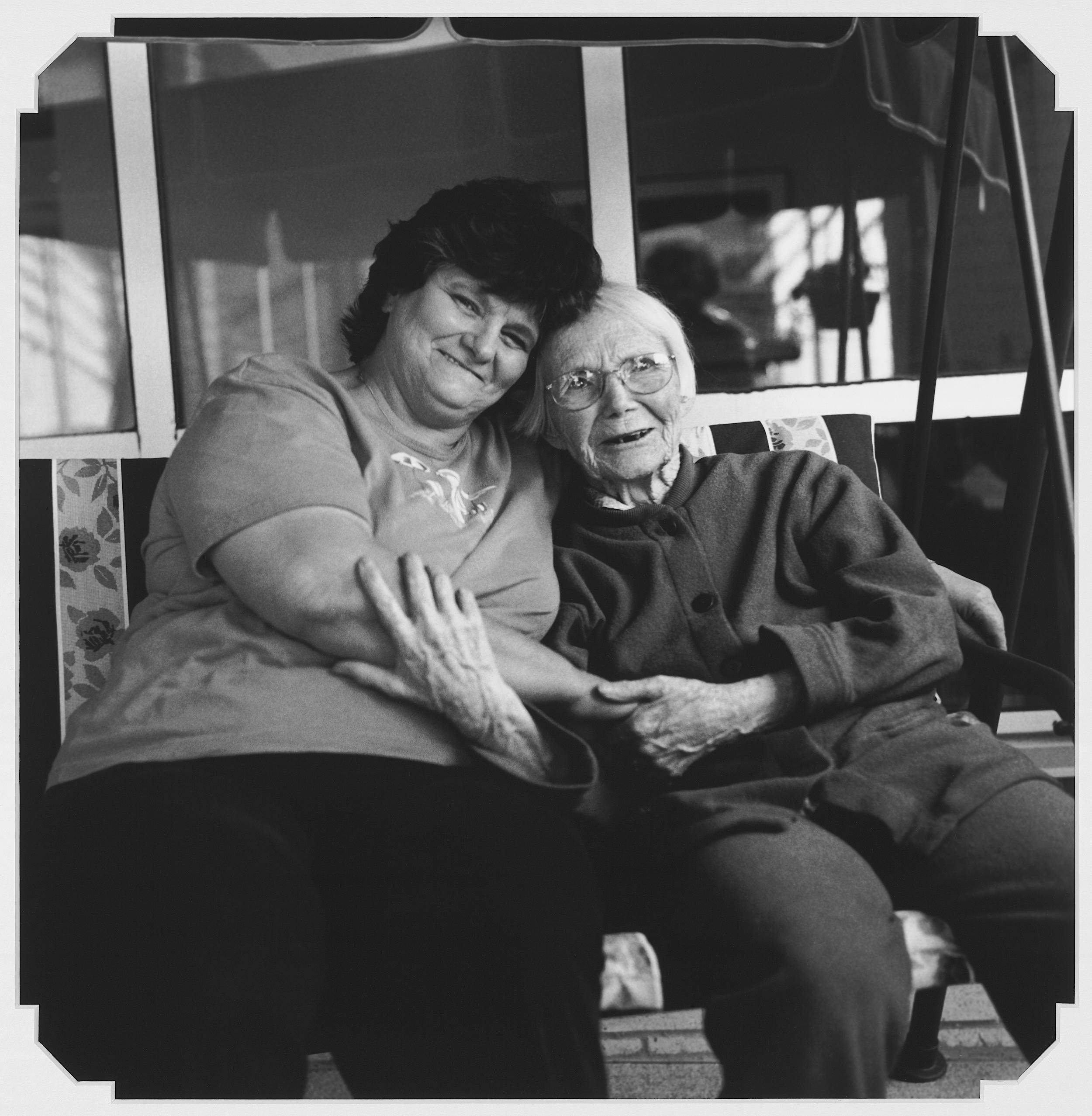 Black and white photo of elderly woman holding hands and arm of younger woman, both wearing warm affectionate smiles.