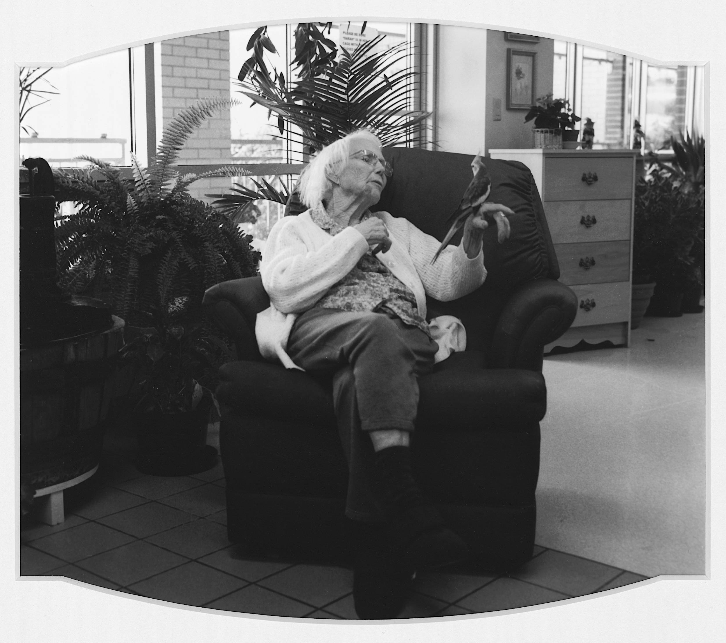Black and white photo of elderly woman seated on sofa chair holding a bird on outstretched hand.