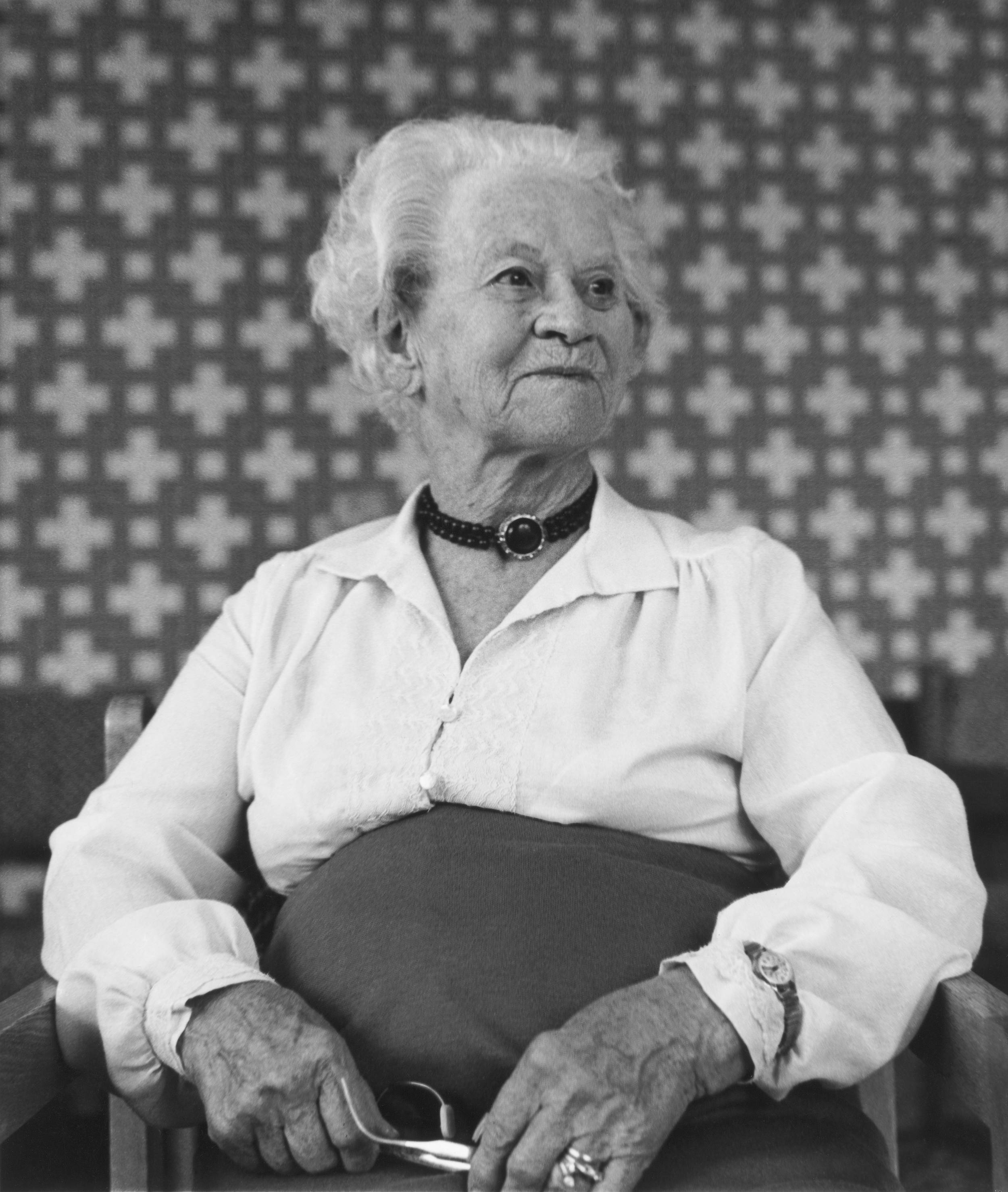 Black and white photo of elderly woman seated with wrist watch on outside of white blouse, looking off to the side very proud and distinguished.