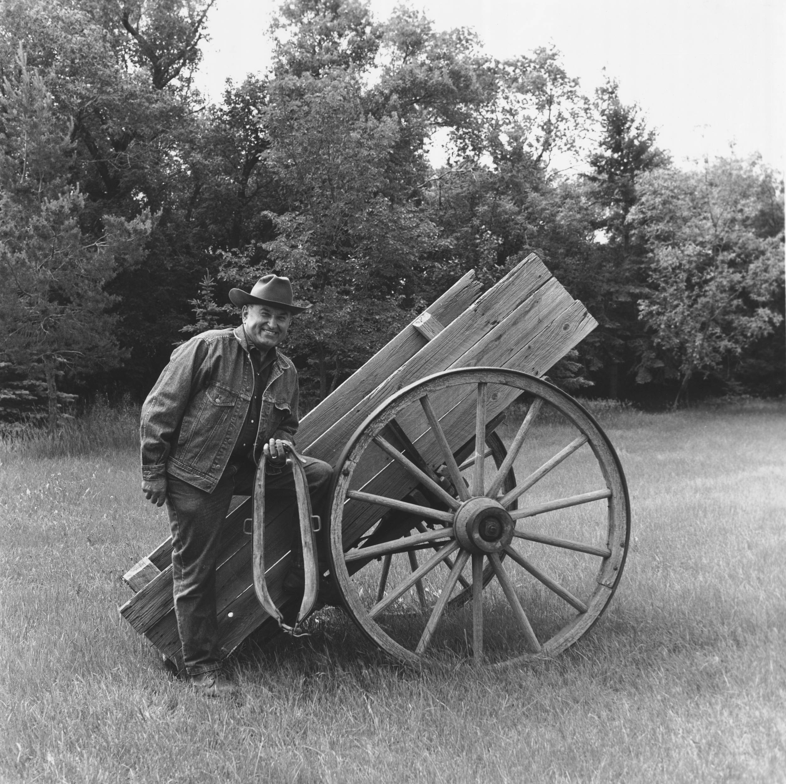 Black and white photo of man standing beside an old wagon cart with large wagon wheel.