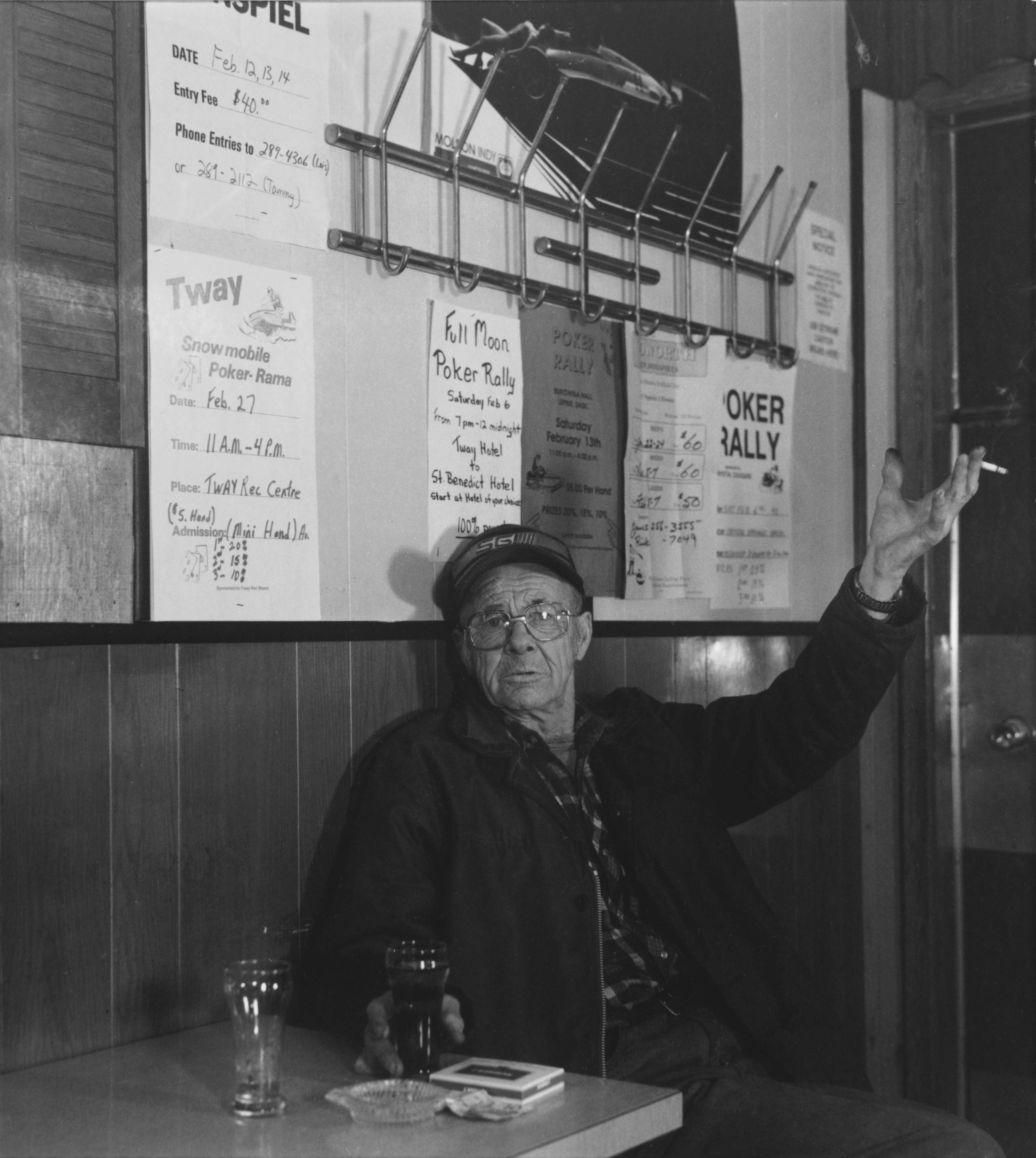 Black and white photo of elderly man inside a rural bar holding up in one hand a cigarette and in the other grasping a beer on the small round table.