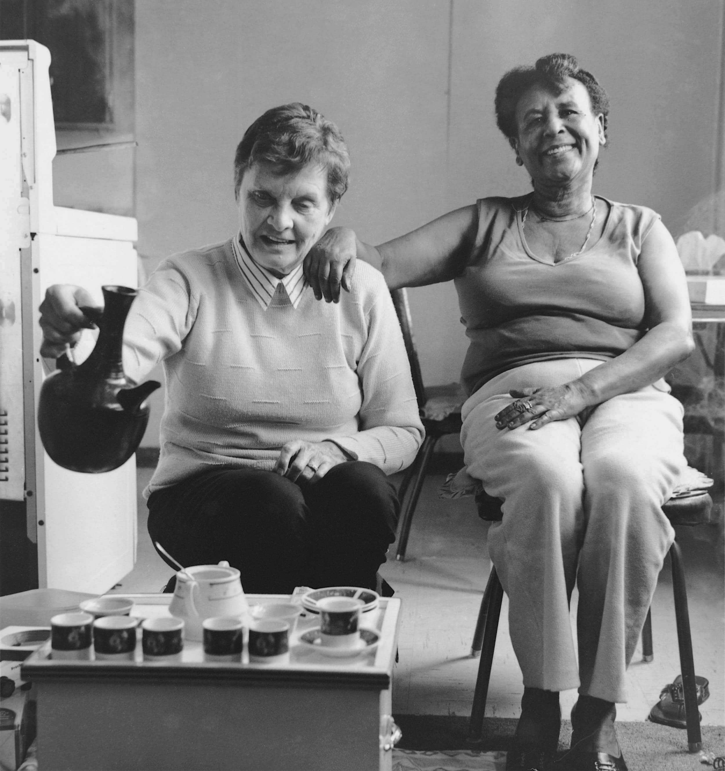 Black and white photo of two woman, both seated and smiling, one pouring coffee from tall Ethiopian coffee container, and the other with outstretched arm around her shoulder.