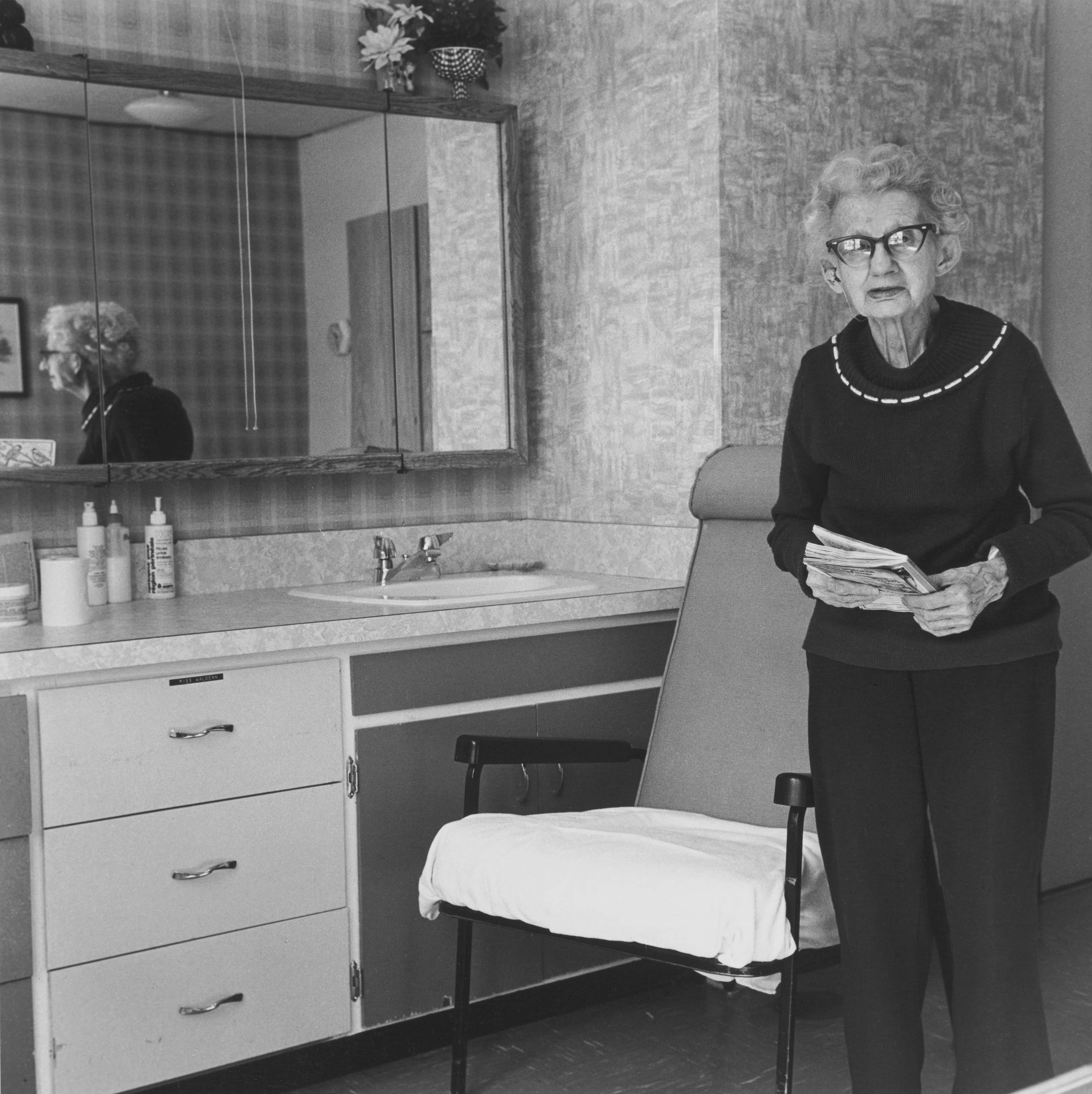 Black and white photo of elderly woman standing holding a book in both hands with her reflection seen in mirror behind her.