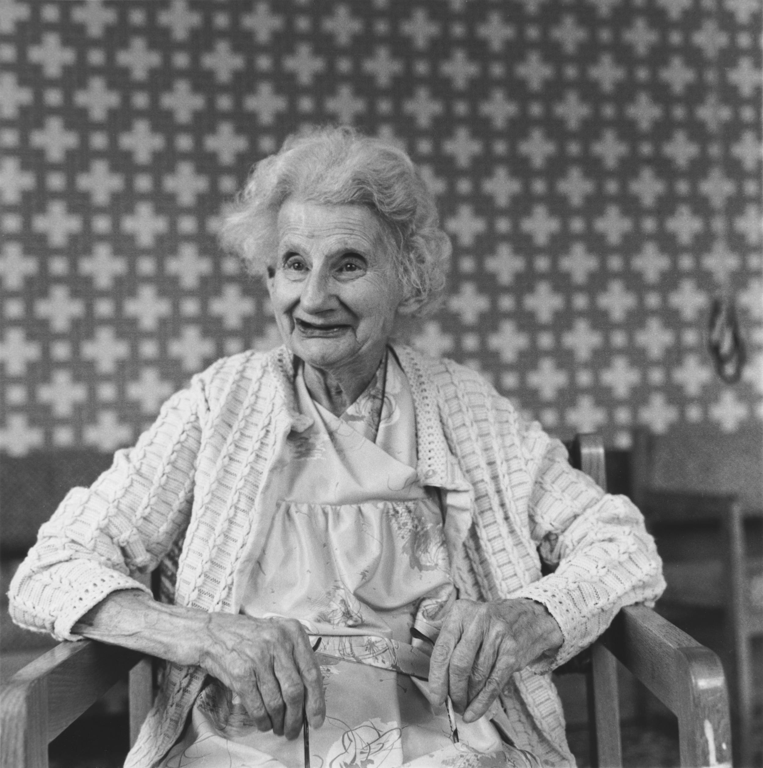 Black and white photo of elderly woman sitting down wearing beautiful warm smile with checker patterned wallpaper in background.