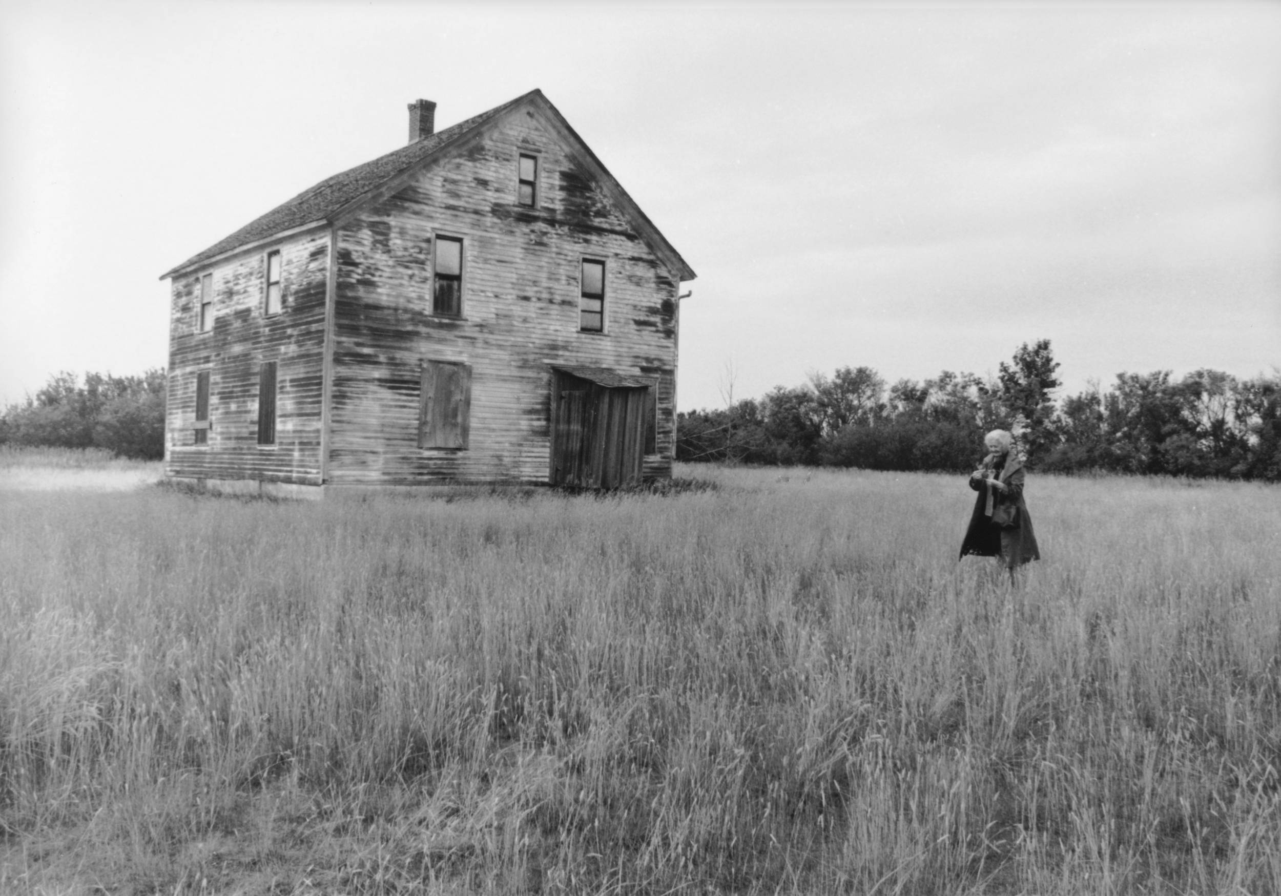 Black and white: wide shot of woman standing in open grass field beside old wooden barn.