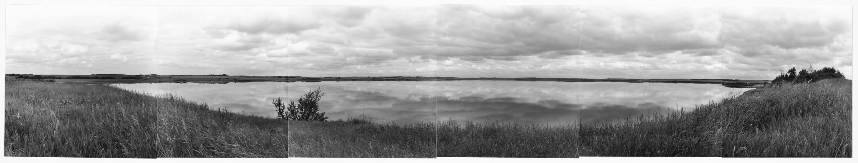 Black and white six panel panorama photo overlooking the lake at Prud’homme. All six panels reveal the beautiful reflection of the prairie sky in the water below.