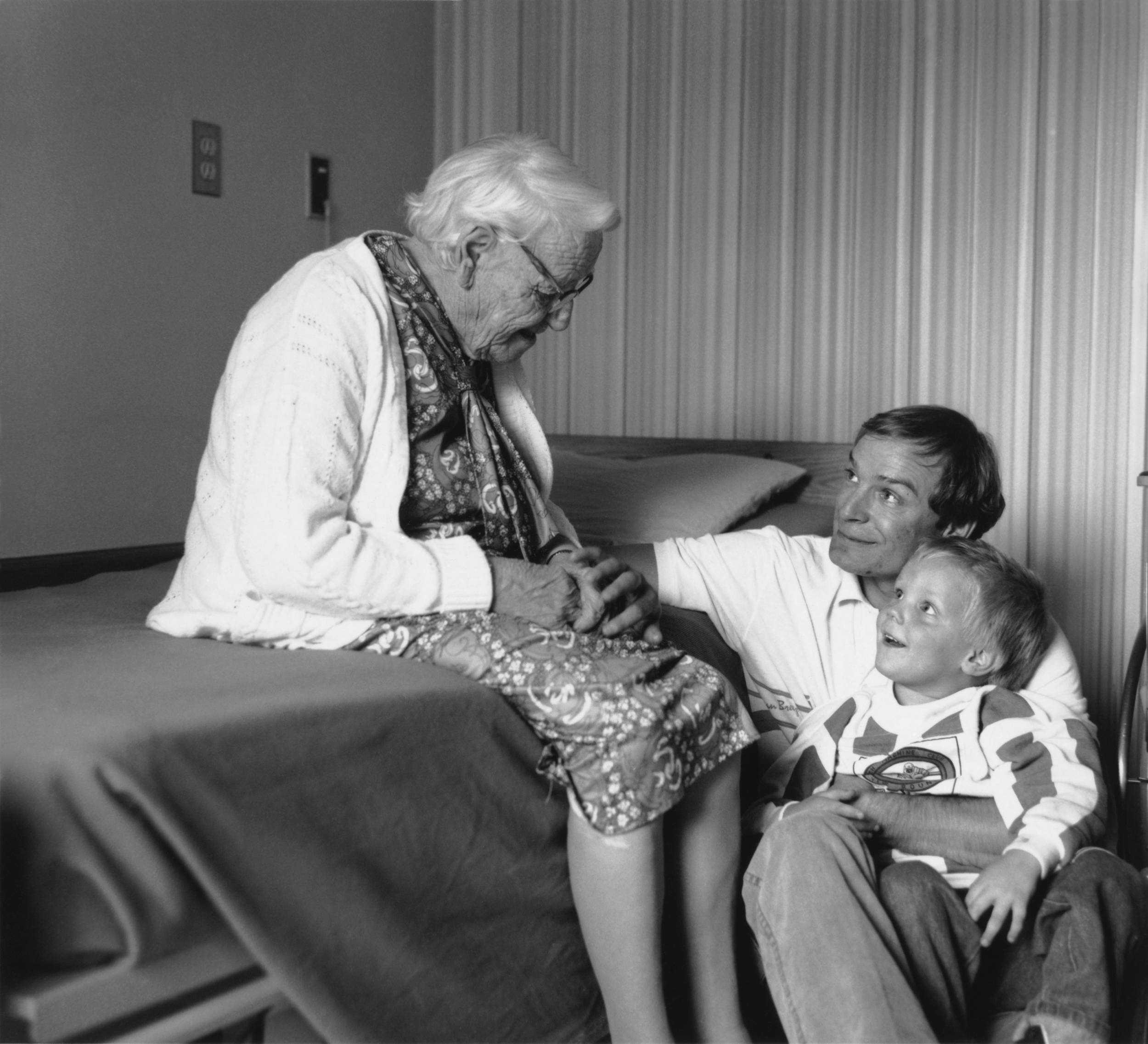Black and white photo of elderly woman seated on bed looking over at son and grandchild who are both looking up at grandma with adoration.