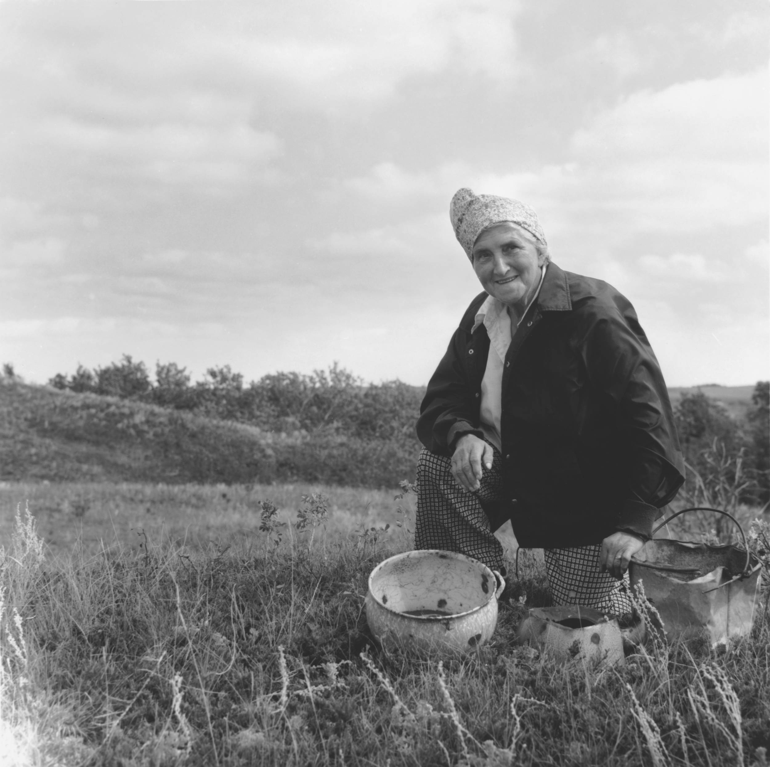 Black and white photo of elderly woman kneeling down in open field with a small pot in front of her. She is wearing a head scarf with warm smile.