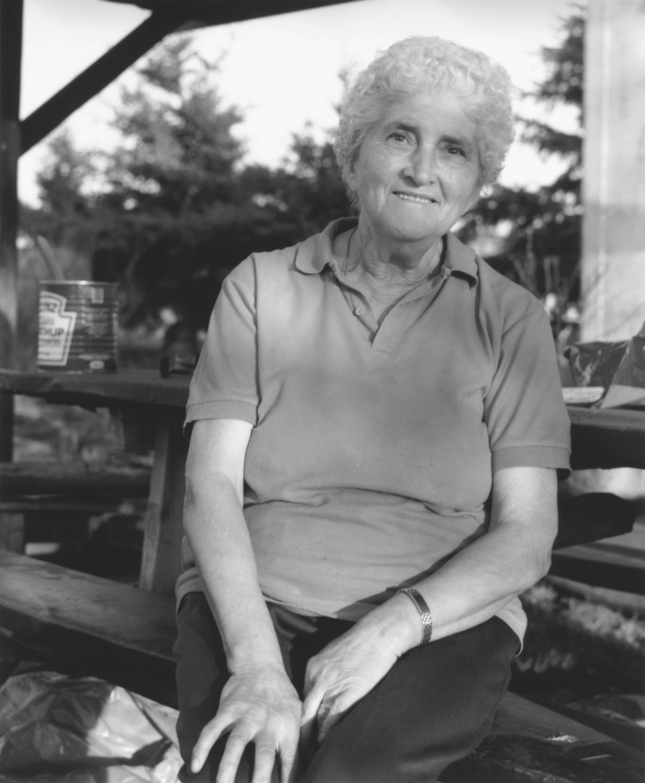 Black and white photo of elderly woman with big smile looking at the camera. The woman is sitting down on a bench with both hands on her knees.