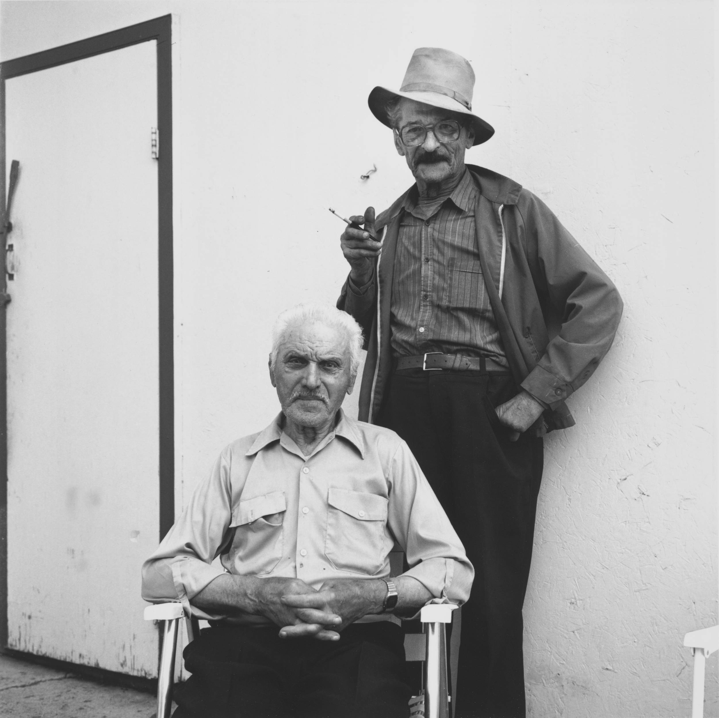 Black and white photo of two elderly men, one seated on lawn chair and other standing behind holding a cigarette in right hand.