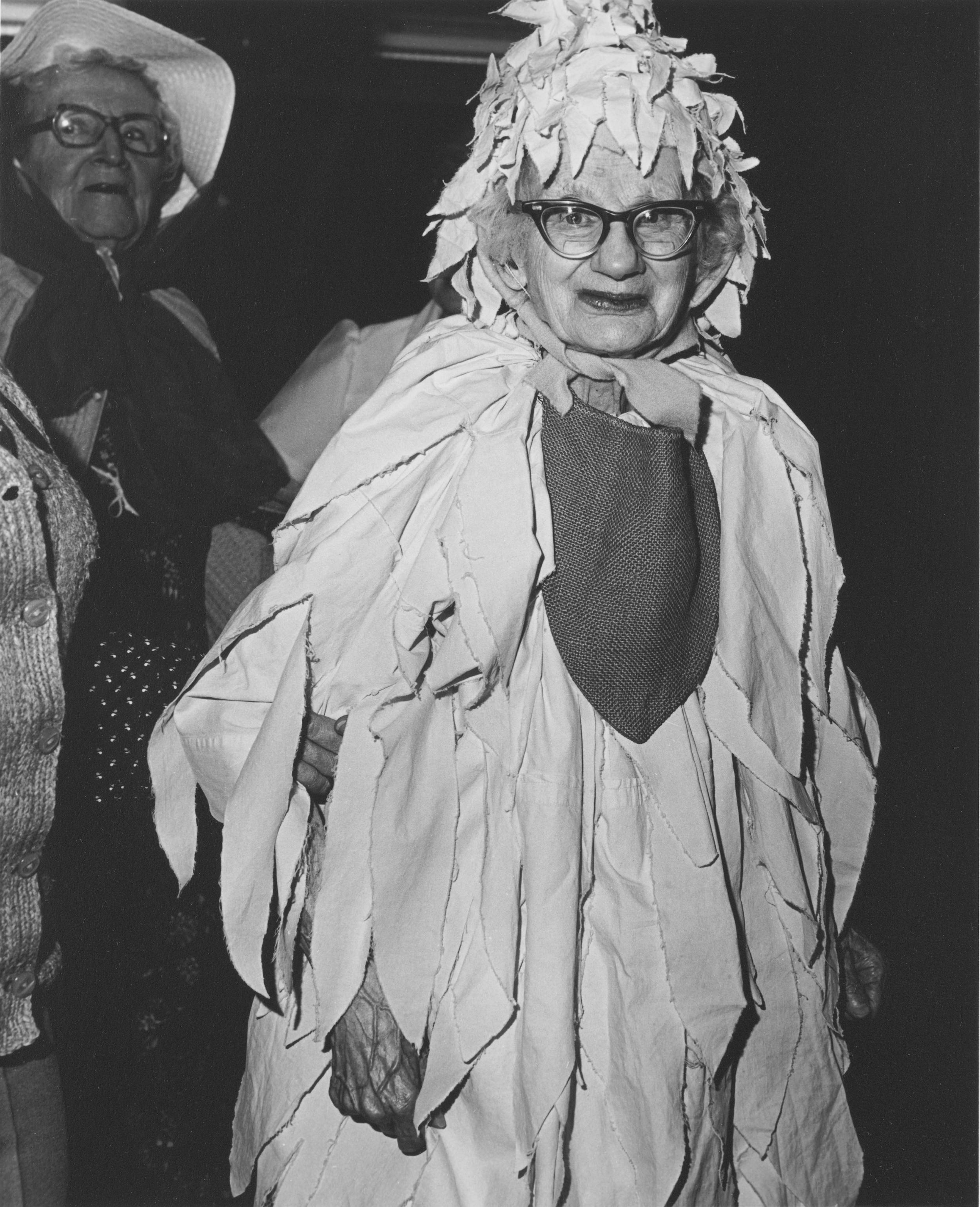 Black and white photo of elderly woman standing, dressed in fancy costume waiting for parade.