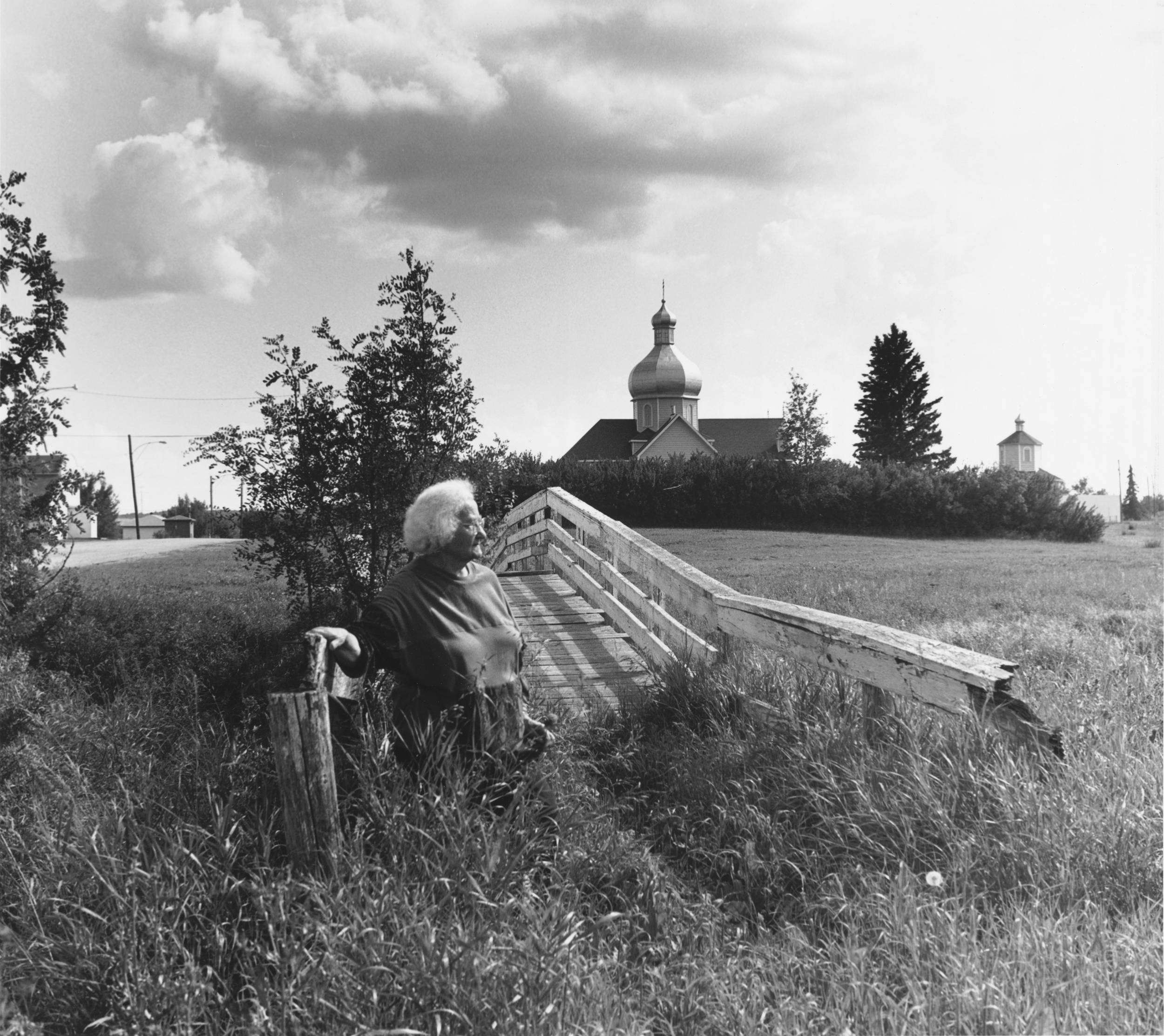 Black and white photo of elderly woman standing sideway at the foot of a small rural bridge, with large orthodox church tower in the background.