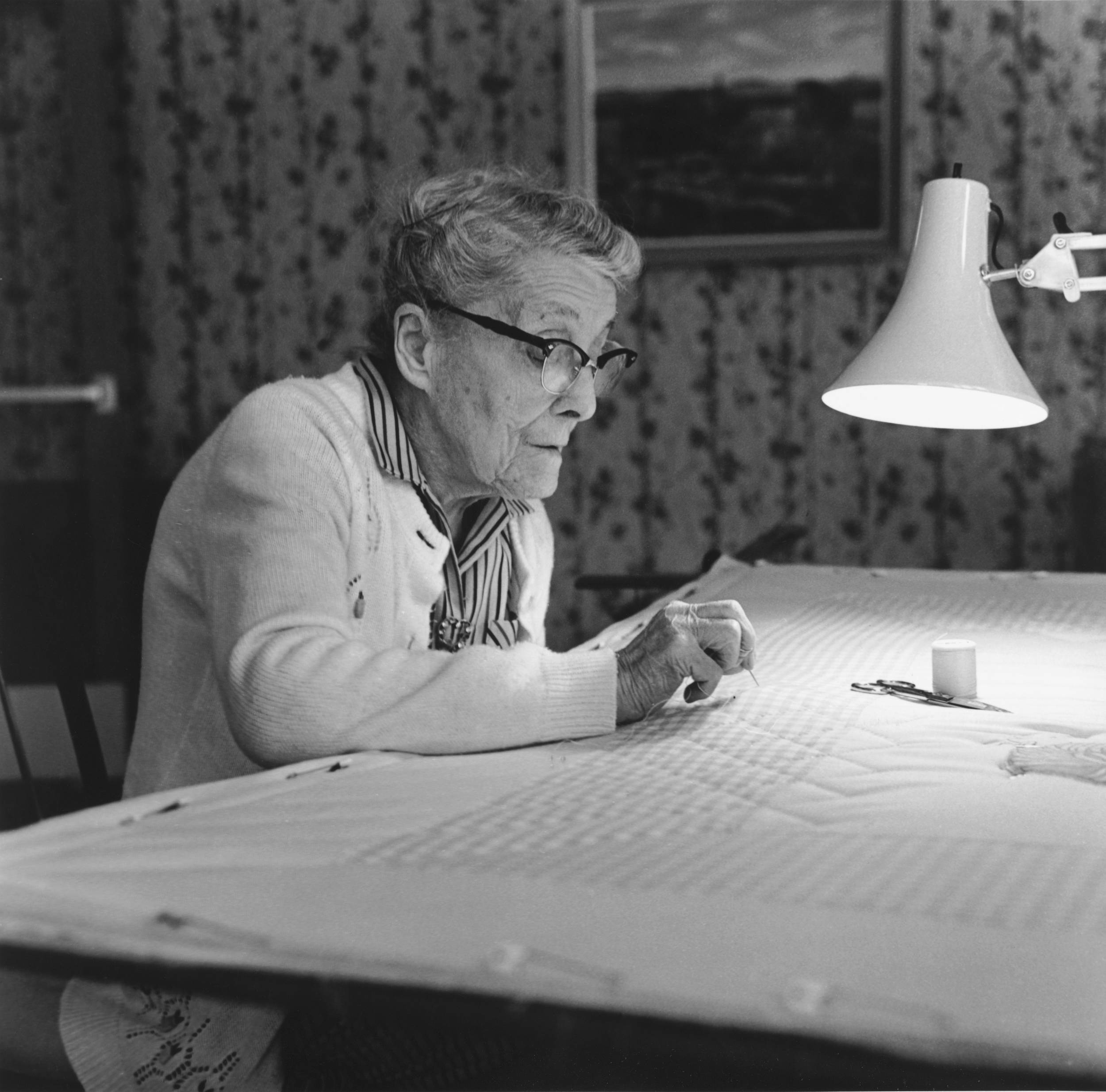 Black and white photo of elderly woman quilting away with needle on table using light from attached extension light.