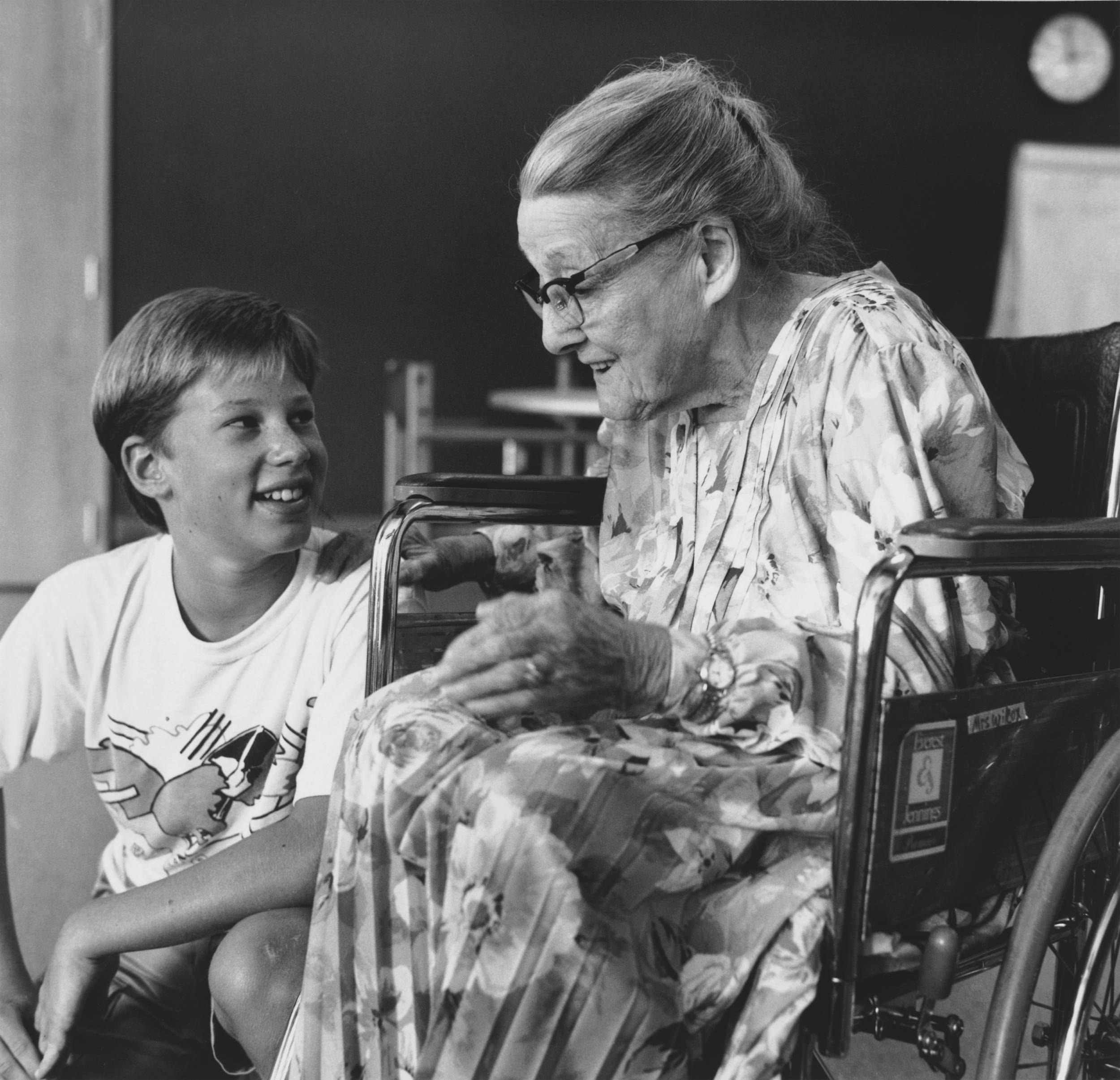 Black and white photo of elderly woman in wheelchair wearing warm smile looking over at young boy.