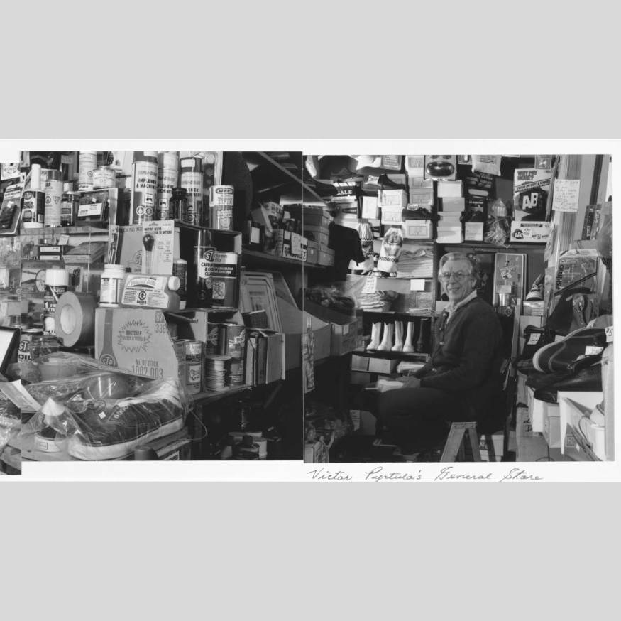 Black and white photo of elderly man standing with arms crossed, very proudly, inside his general store. He is surrounded by all the items which his small store sells.