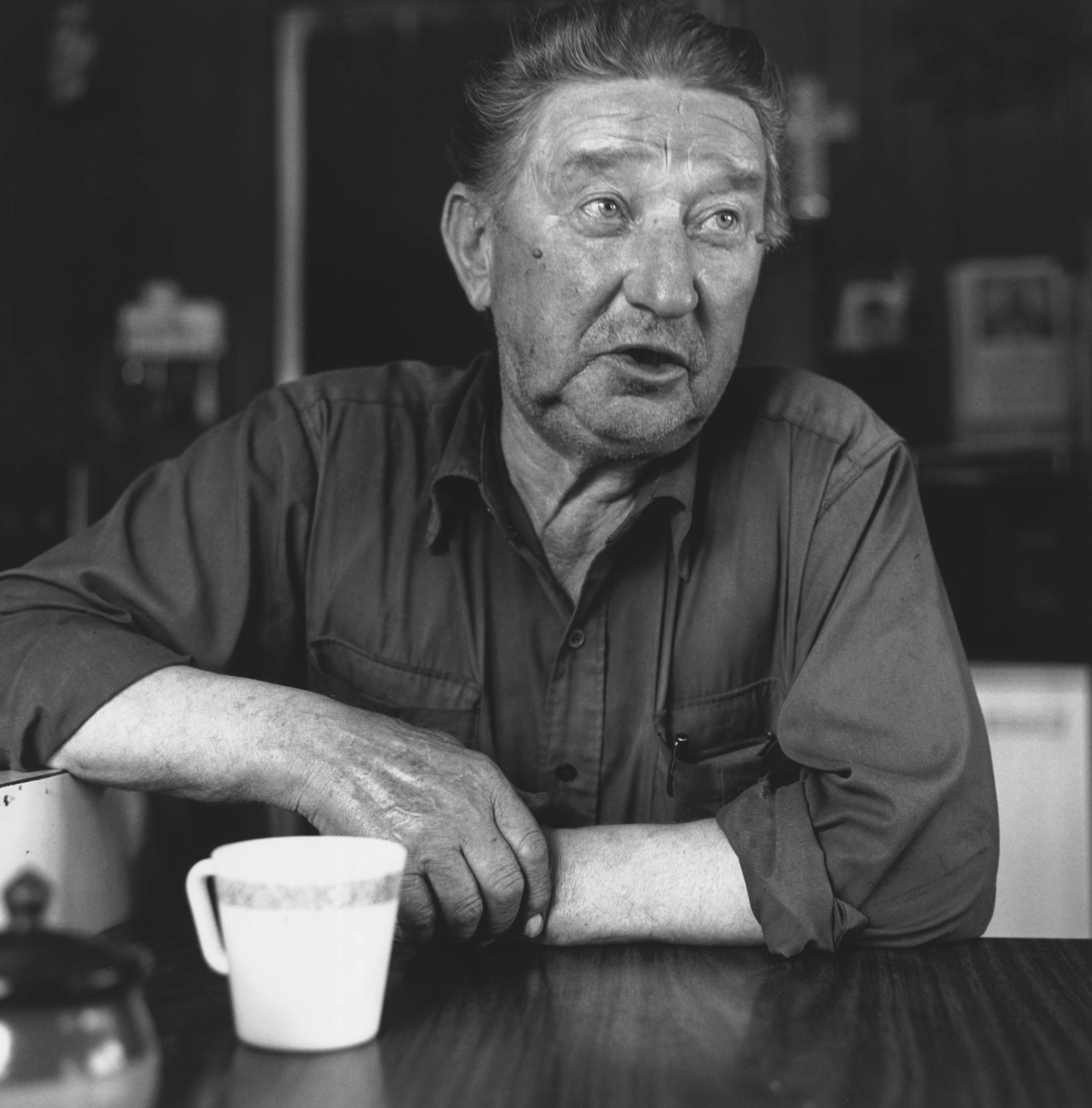 Black and white photo from across the table of an elderly man with coffee cup in front of him.
