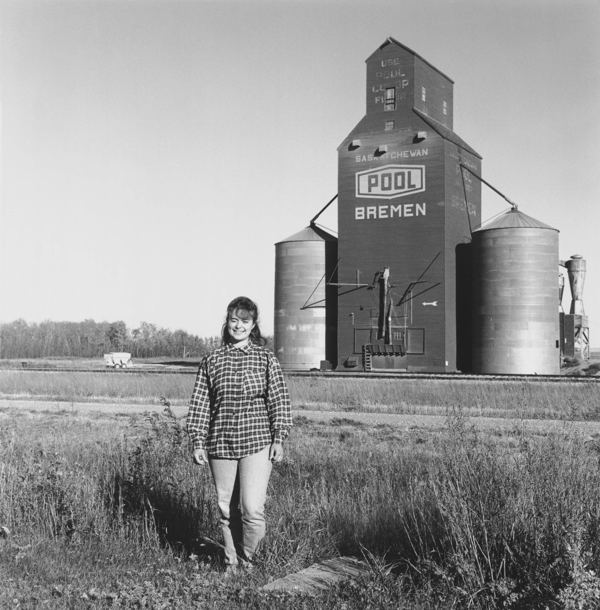 Black and white photo of young woman standing in front of her small-town rural grain elevator. The woman is wearing a big smile and looks very happy standing in her field.