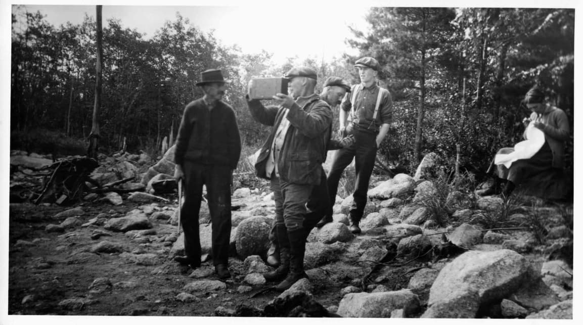 Very old black and white photo of an elderly man among some large rocks holding an old camera.