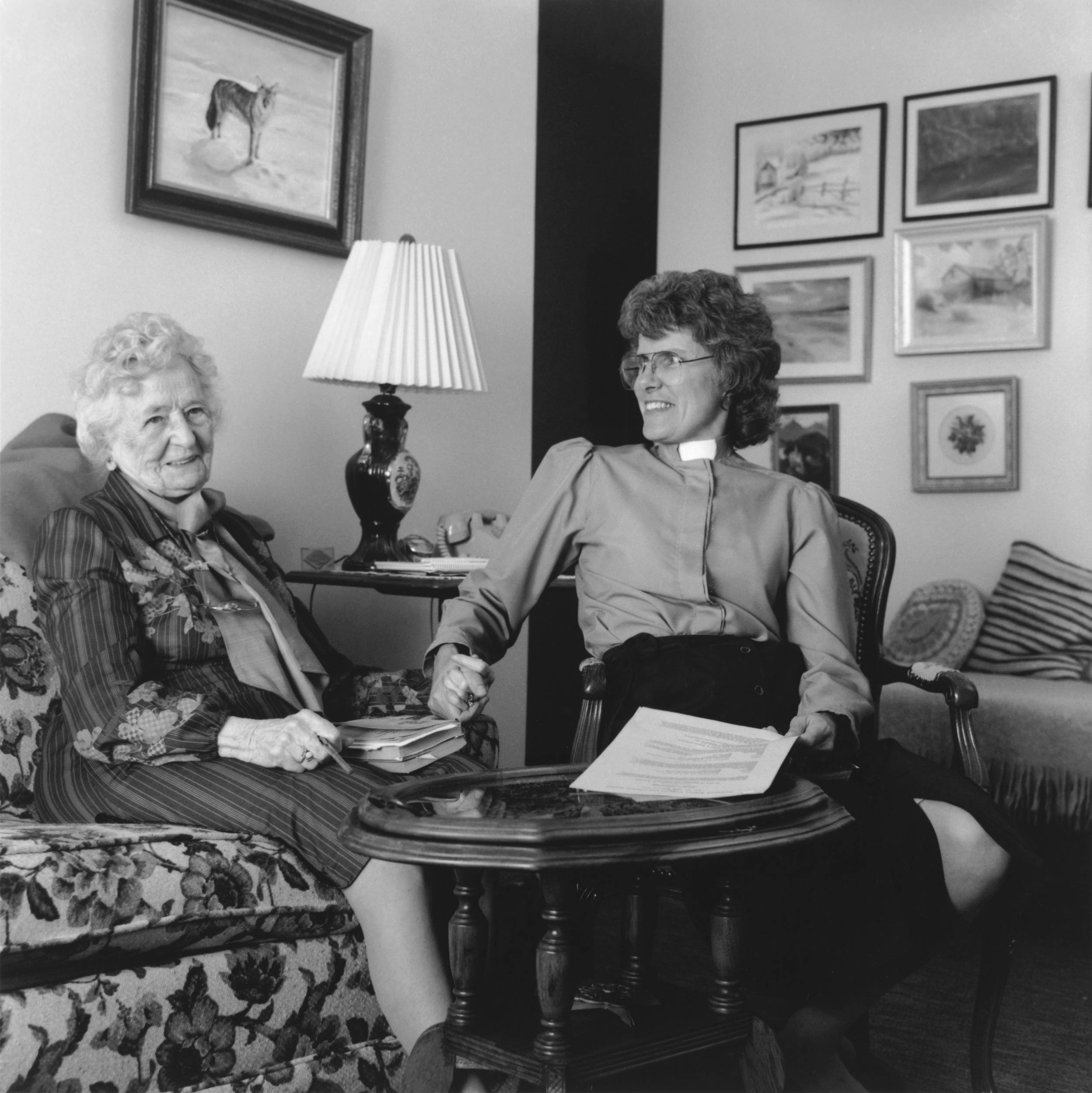 Black and white photo of elderly woman and younger woman seated holding hands. First woman with warm smile and second woman (sister of the church) looking over smiling.