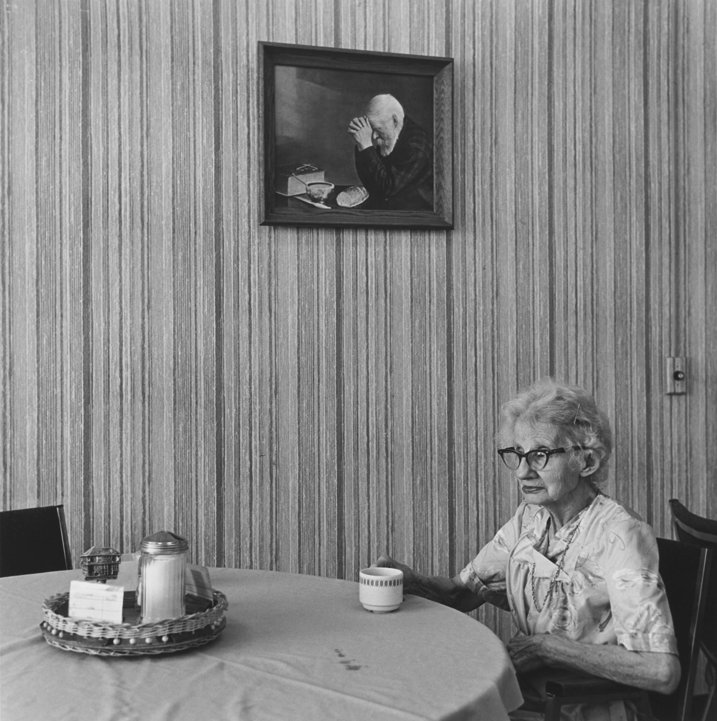Black and white photo of elderly woman in thought seated holding coffee cup on the side of round table, and behind her picture on wall of elderly man praying.