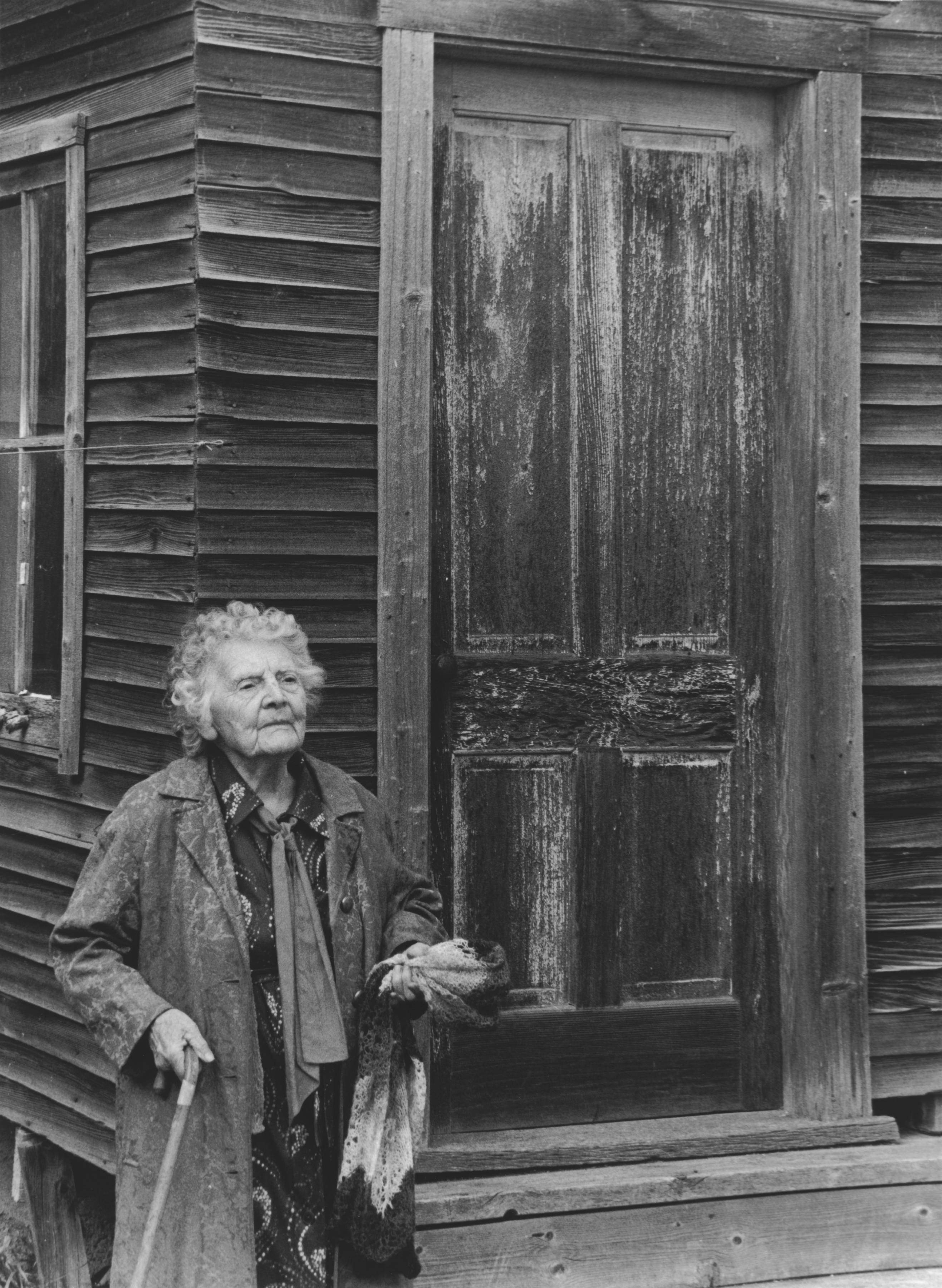 Black and white photo of elderly woman standing strong in front of small old wooden barn, holding scarf in one hand and cane in the other.