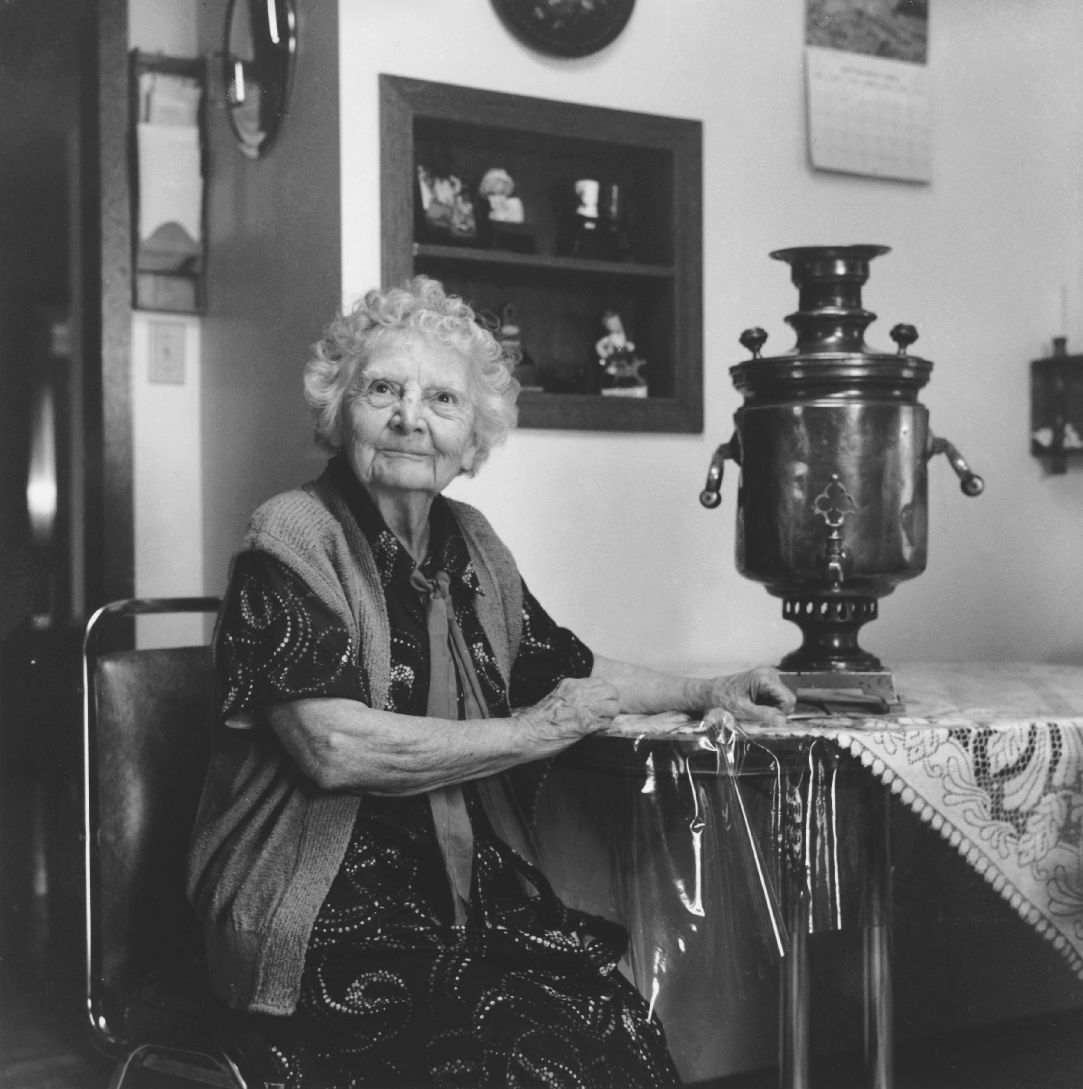 Black and white photo of elderly lady sitting beside her family Samovar, with both hands close by.