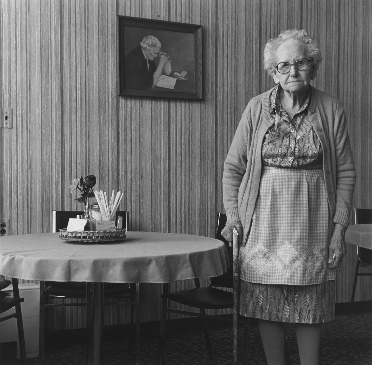 Black and white photo of elderly woman with cane standing beside small dining table, striped wallpaper background