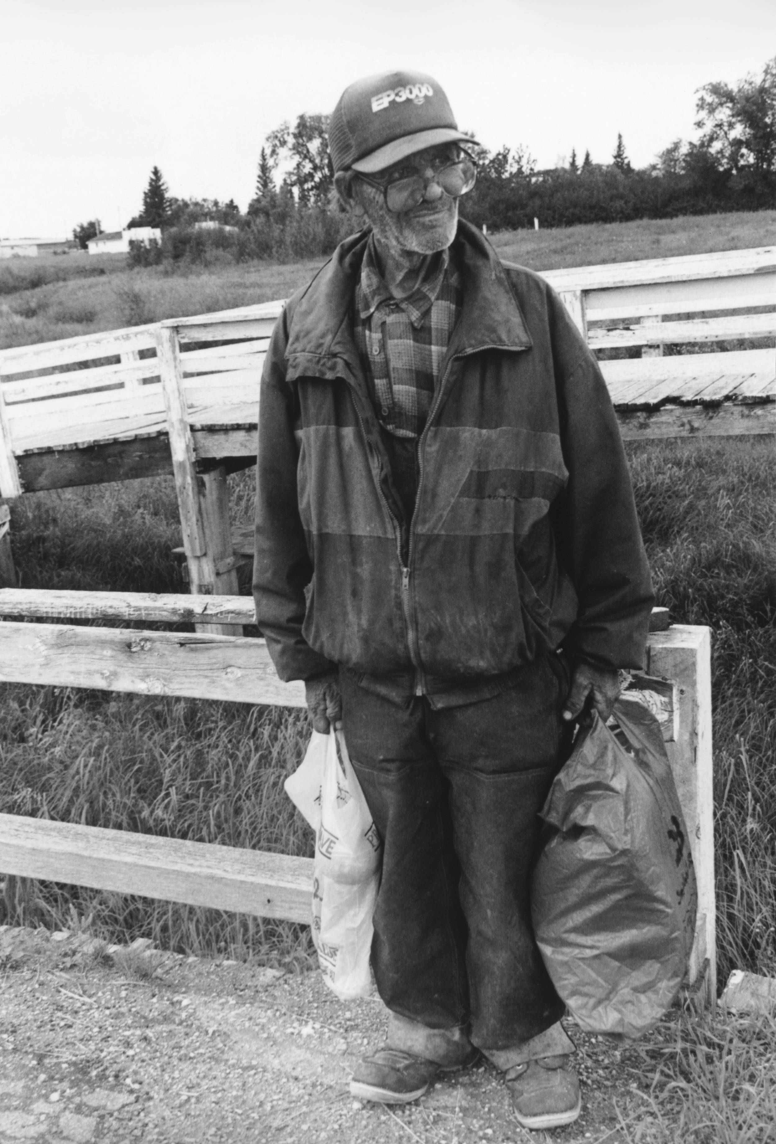 Black and white photo of elderly man outside carrying in both hands two large bags. The man has on big glasses, an old hat, worn coat and pants and large old shoes.