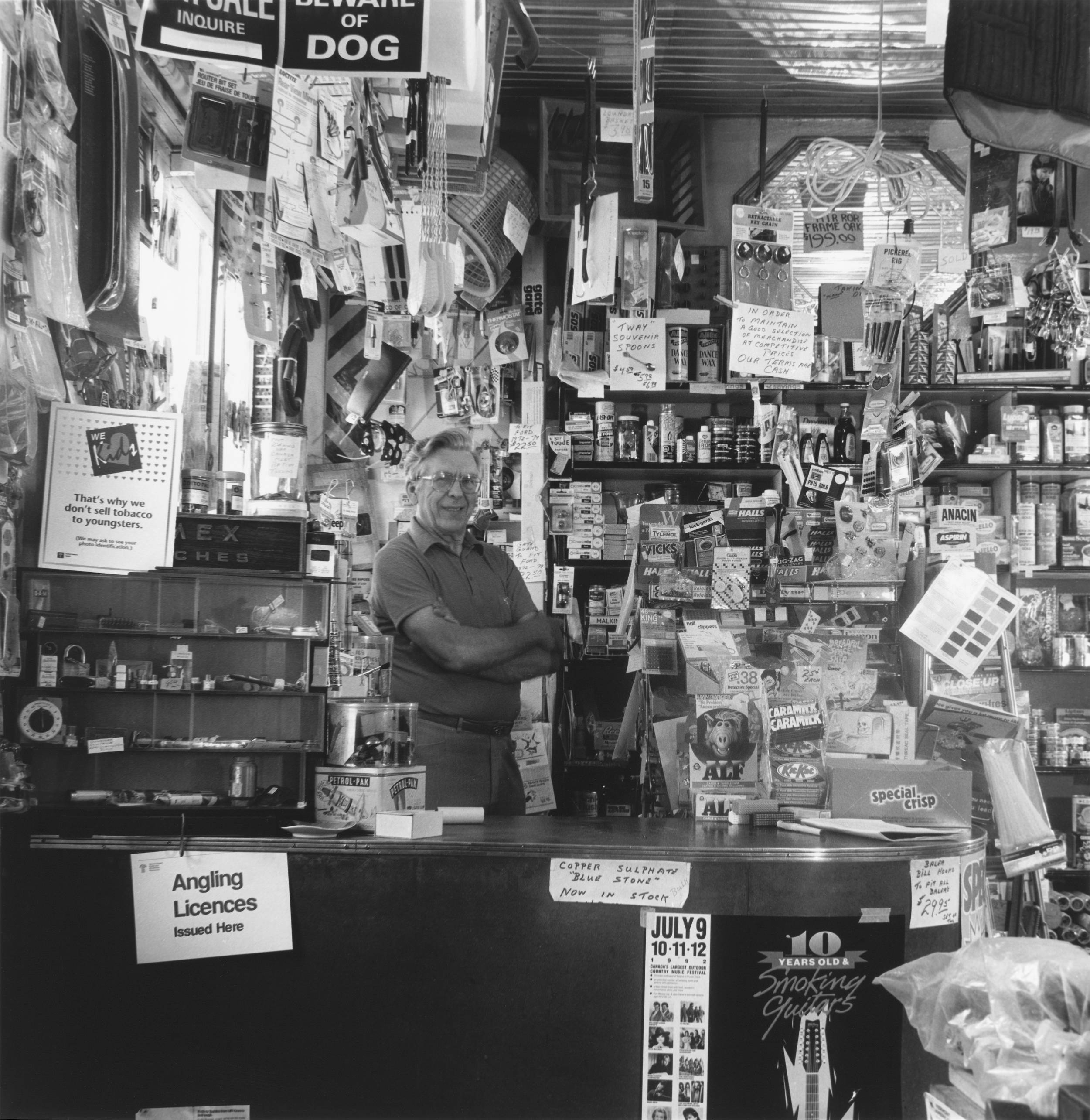Black and white photo of elderly man standing with arms crossed, very proudly, inside his general store. He is surrounded by all the items which his small store sells.